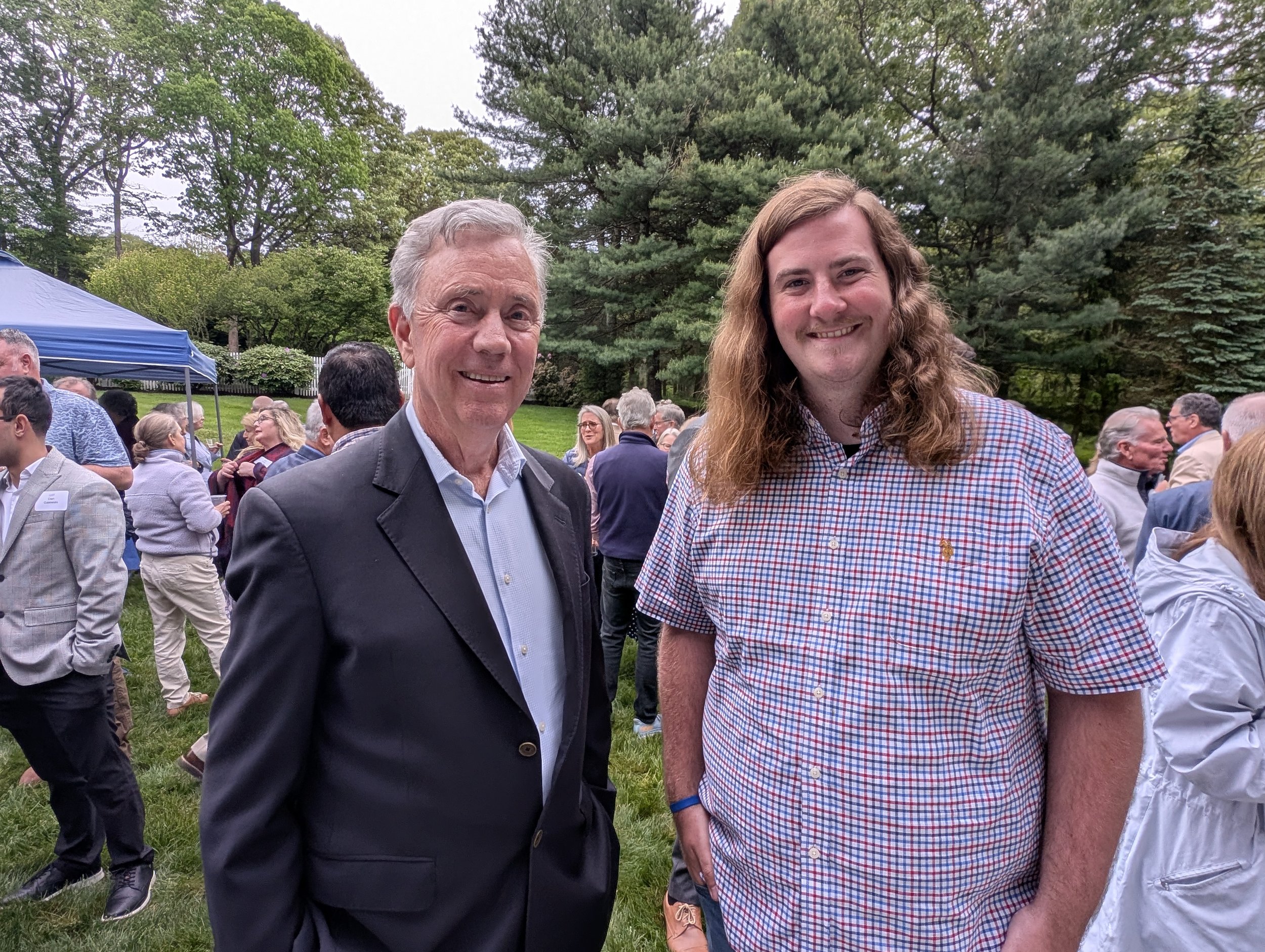 Two men smiling at an outdoor event with many people gathered around, trees in the background, and a blue canopy tent.