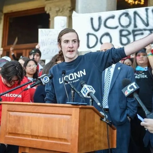 A man wearing a blue UConn sweatshirt speaking at a podium during a protest or rally. Behind her, people are holding signs, one reads 'UCONN DO BETTER' and another partially visible. Several microphones are positioned at the podium.