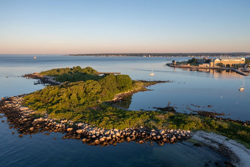 Aerial view of a small, tree-covered island with rocks along its shoreline, surrounded by calm water with boats, and a distant cityscape on the horizon during sunset.