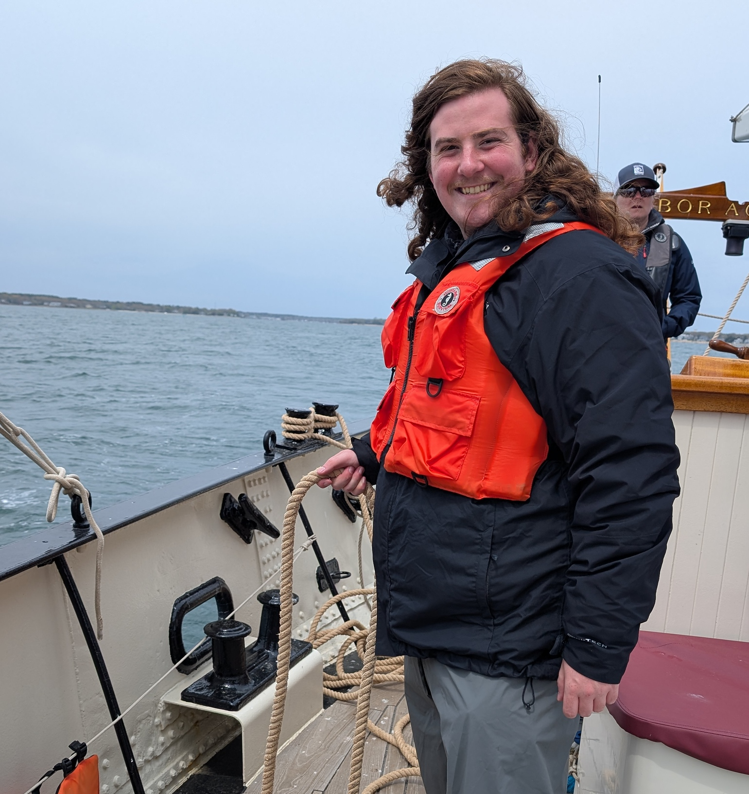 A man with long curly hair smiling on a boat, holding a rope, with another person in the background and a body of water in the distance.