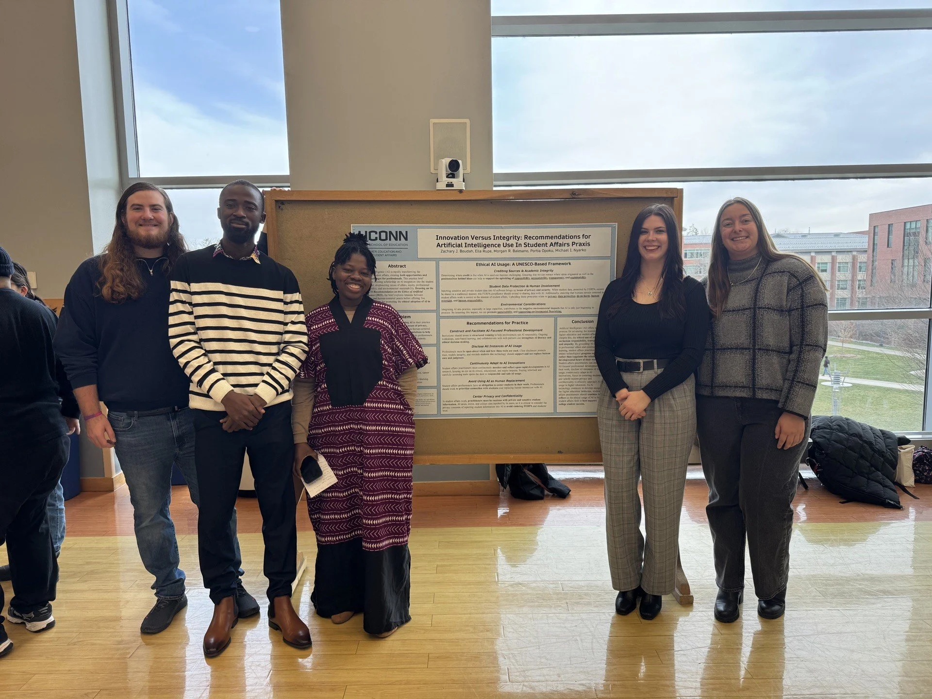 A group of six diverse students standing in front of a research poster at an academic event, with a window and outside view in the background. They are smiling and dressed in casual and business casual attire.