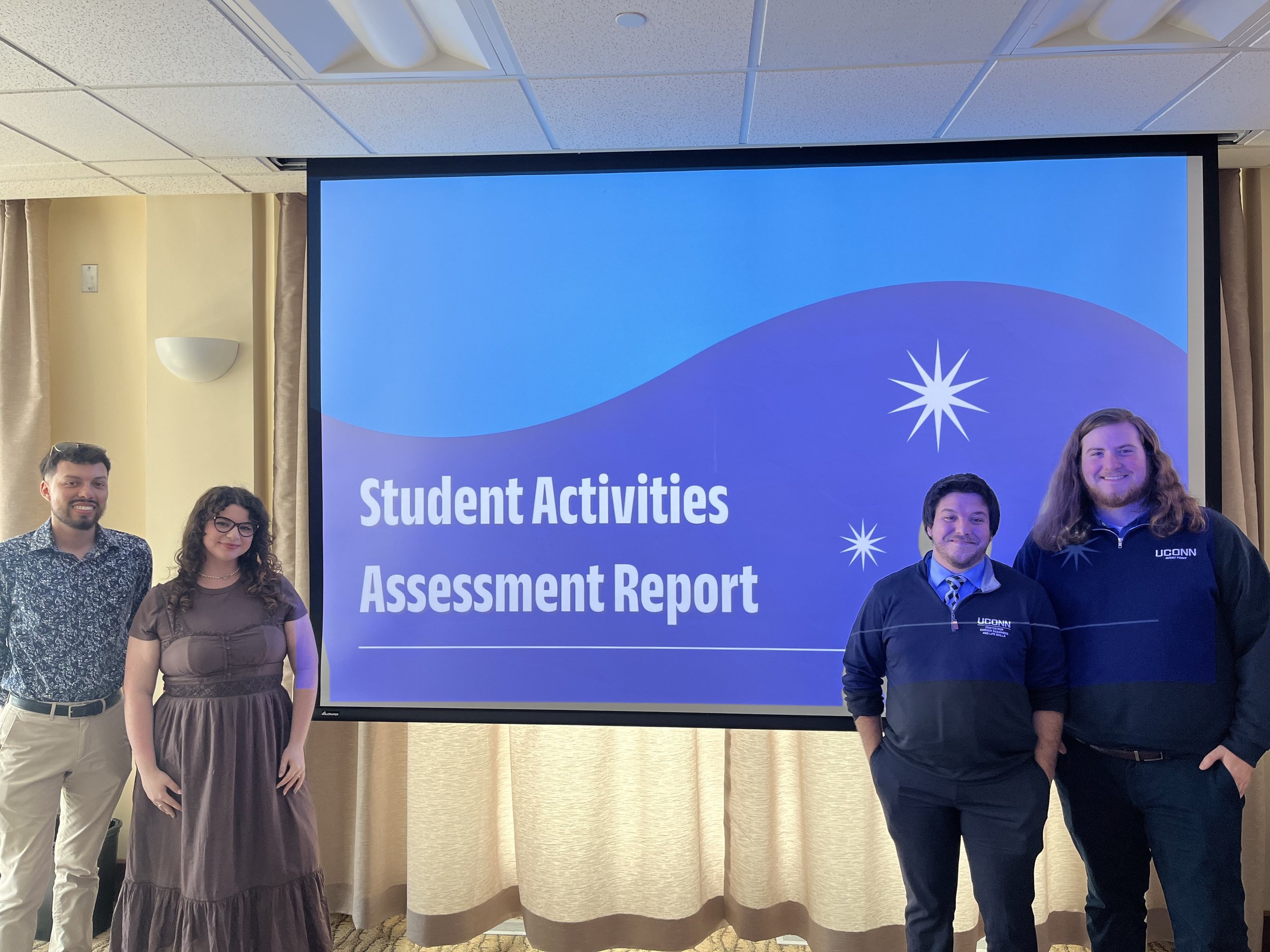 Group of five young adults standing in front of a large screen displaying a presentation titled "Student Activities and Assessment Report." The group includes two women and three men, smiling for the photo. The setting appears to be a conference room or classroom with beige curtains and ceiling lights.