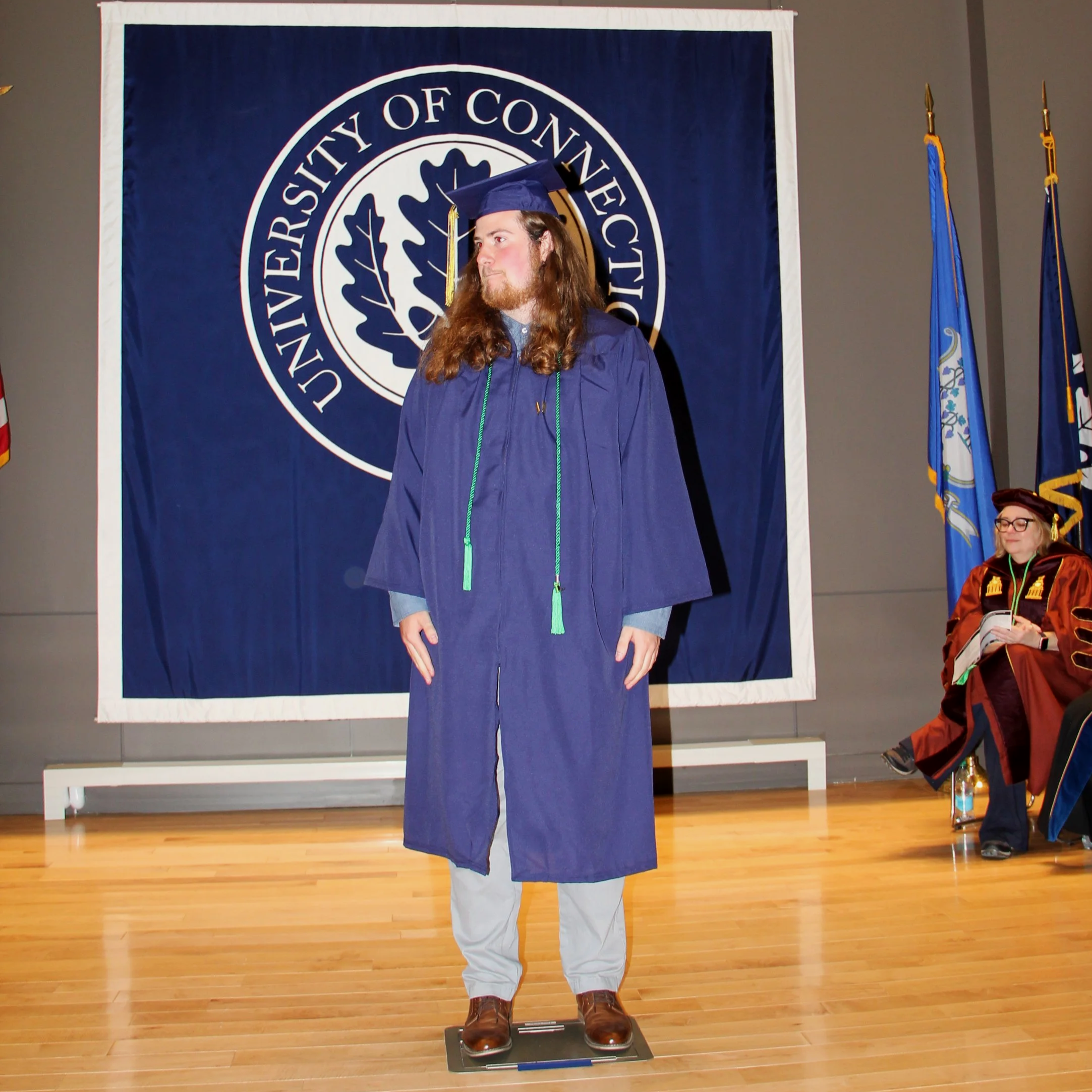 A person in a blue graduation gown and cap stands on a scale, with a large blue and white banner of the University of Connecticut in the background. To the right, a person in academic regalia is seated, and flags are displayed.