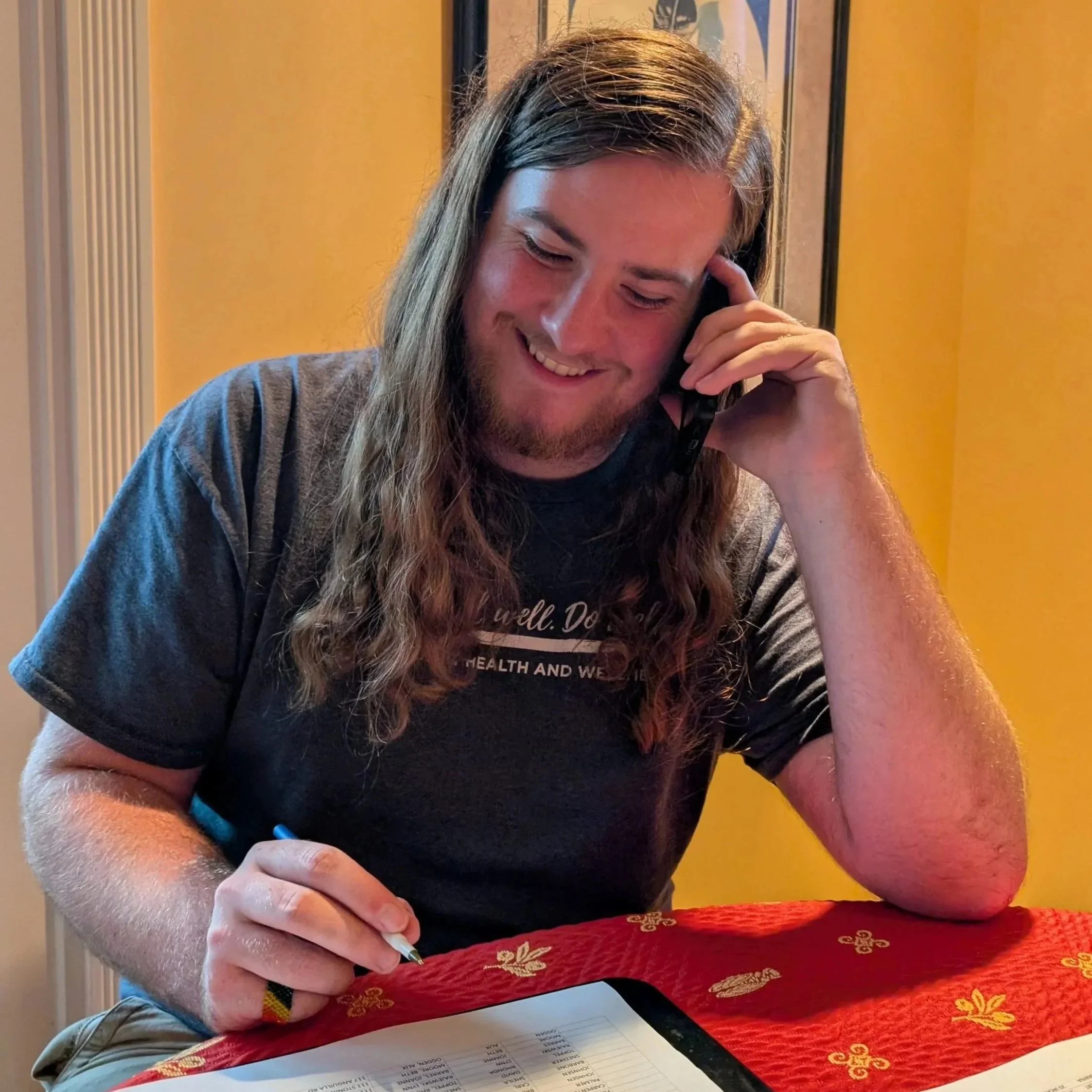A woman with long brown hair, smiling, talking on a black phone while signing a document at a table covered with a red decorative cloth with gold floral patterns.