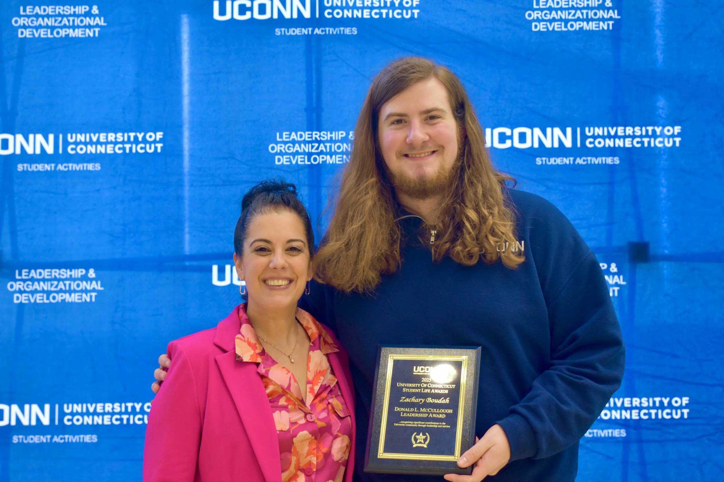 Two smiling women standing in front of a blue UConn banner. One woman is wearing a pink blazer and floral blouse, the other is in a dark jacket holding a framed award. The background includes the university's name and phrases related to student activities and leadership.