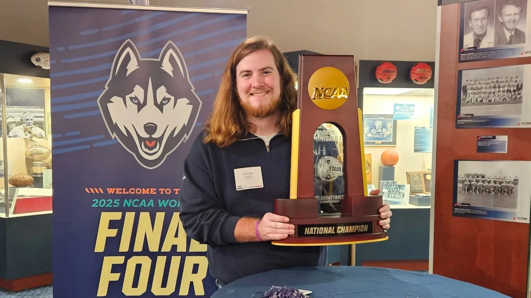 A young man with long hair and a beard holding a large trophy that says 'National Champion' at the 2025 NCAA Final Four event. He is smiling and standing in front of a large banner with a wolf logo and the text 'Final Four'. The background includes display cases with sports memorabilia and photos of basketball teams.