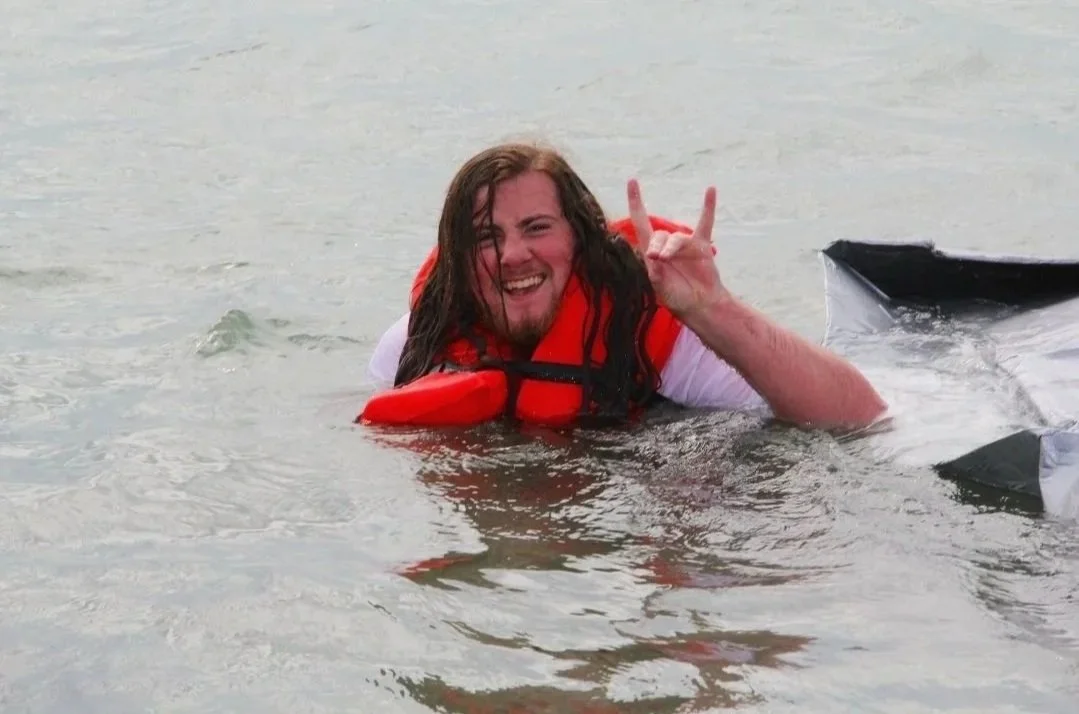 A man with long brown hair, wearing a white shirt and a red life jacket, smiling and making a peace sign while floating in water near a boat at UConn Avery Point Cardboard Boat Race