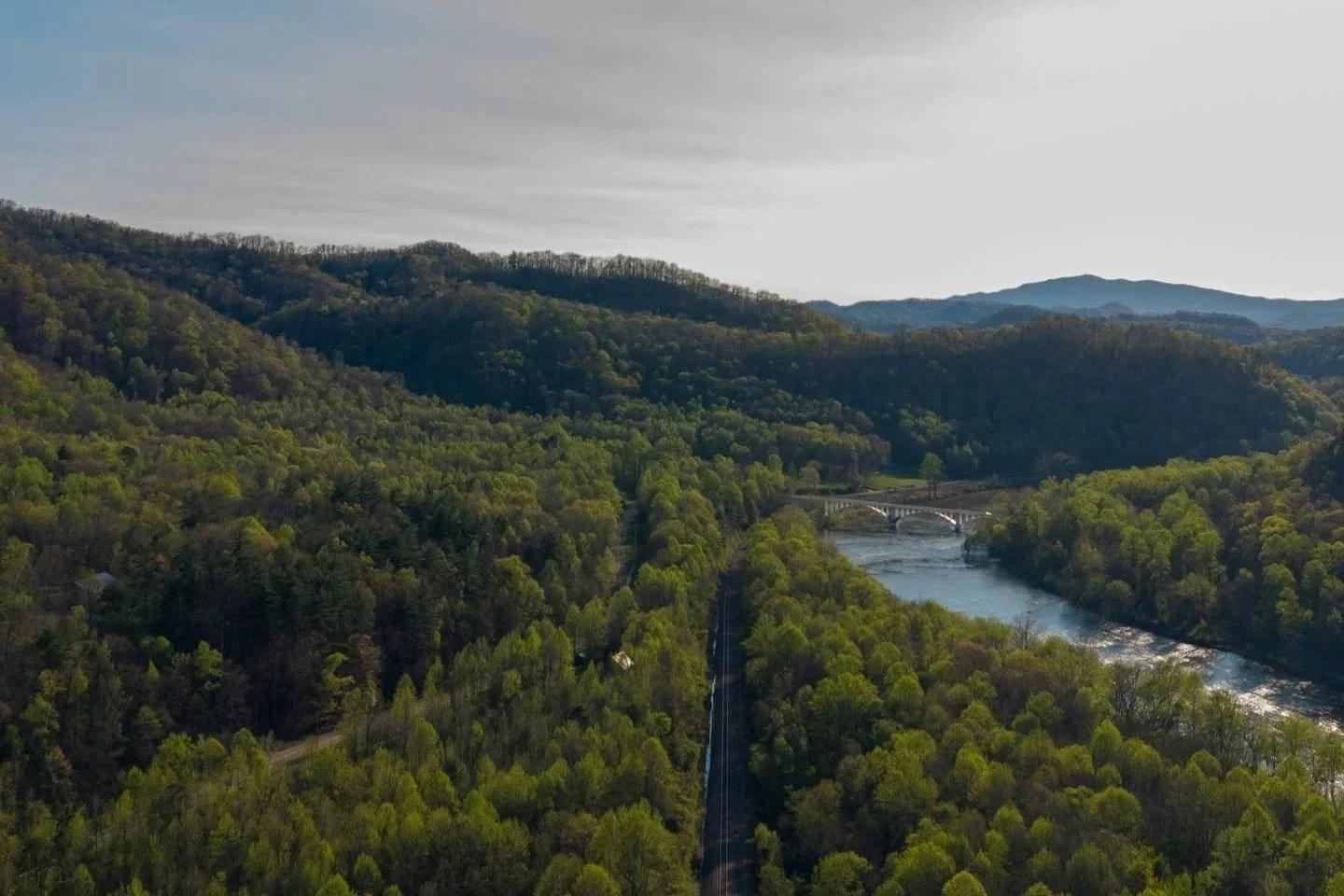 A scenic view of a river flowing through a forested valley with mountains in the background, under a partly cloudy sky.