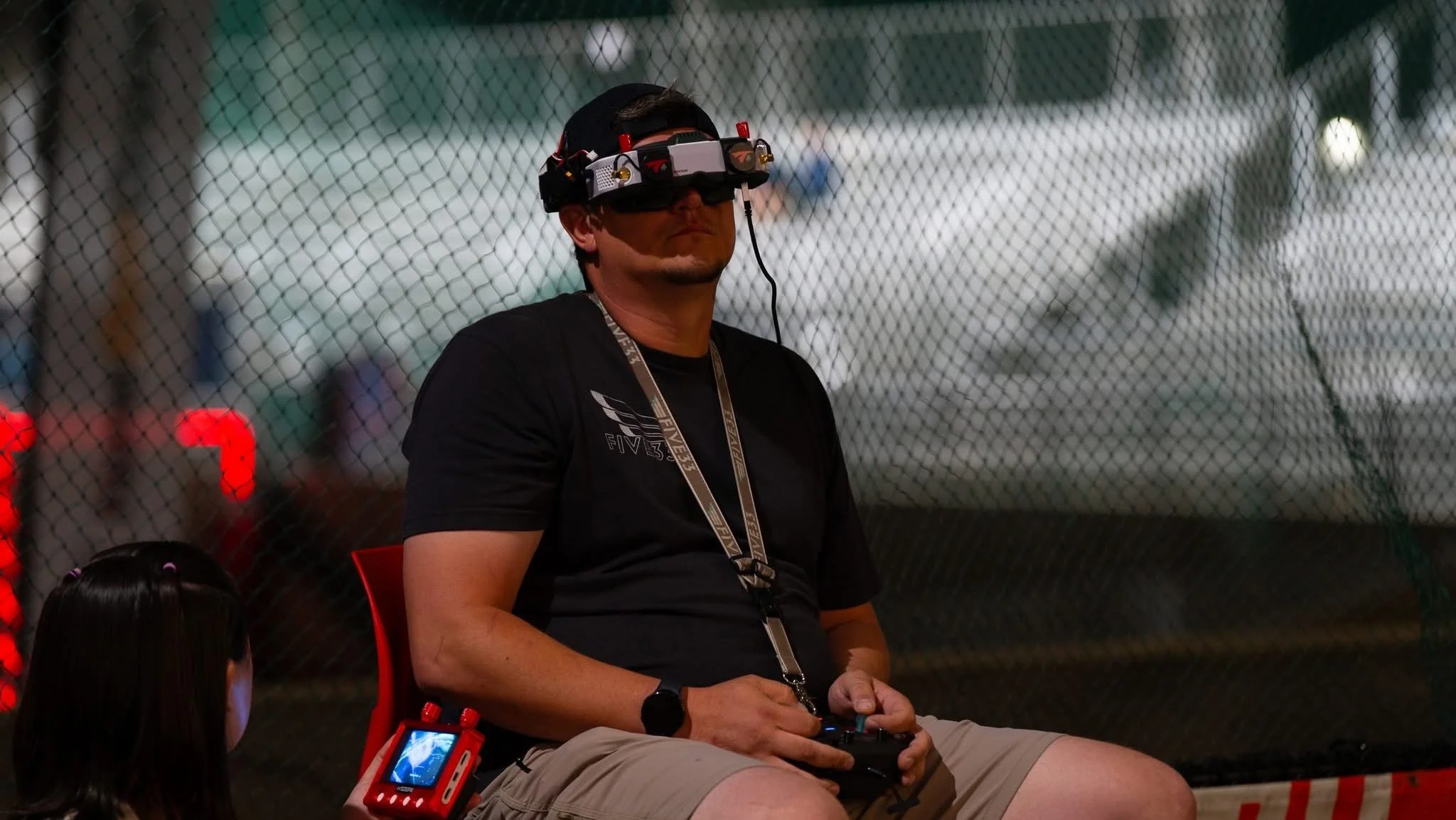 Man wearing virtual reality headset and sitting in front of a chain-link fence, with children nearby.