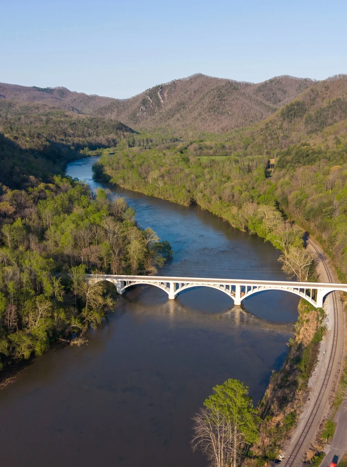 An aerial view of a river winding through a forested valley with mountains in the background. A white bridge spans the river and a railroad track runs alongside the river on the right side of the image.