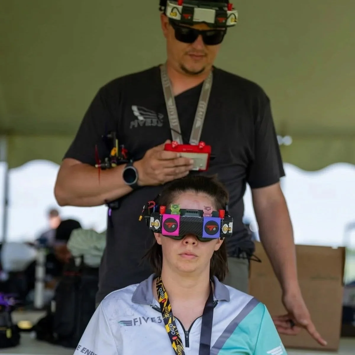 A woman wearing a multi-colored drone racing headset and a lanyard, with a man standing behind her. The man is wearing sunglasses, a headset with a camera, and a black shirt. They appear to be in a tent or covered outdoor area.