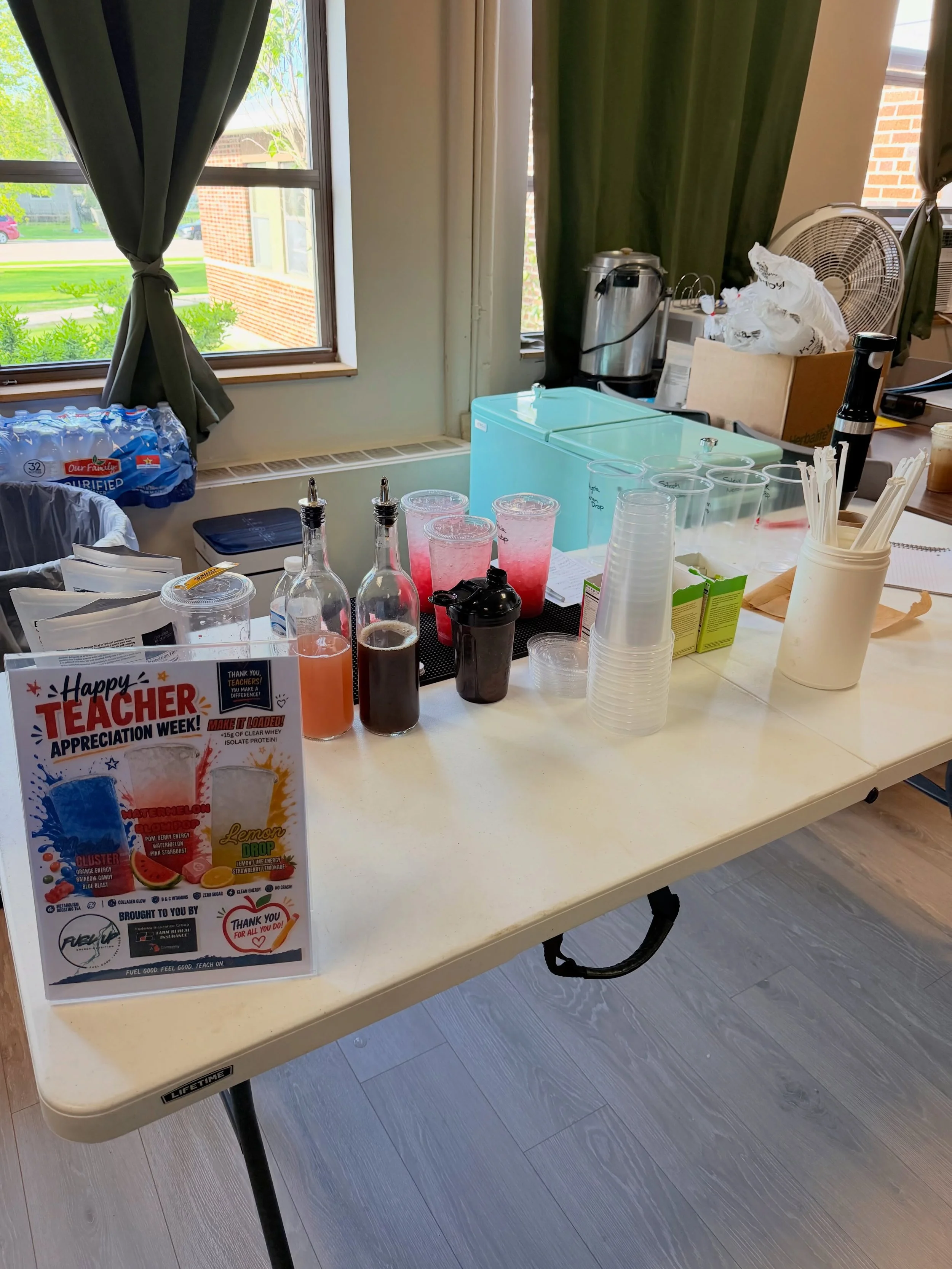 A table with various drinks, cups, and snacks, next to a window with green curtains, and a promotional flyer about energy drinks and teacher appreciation.