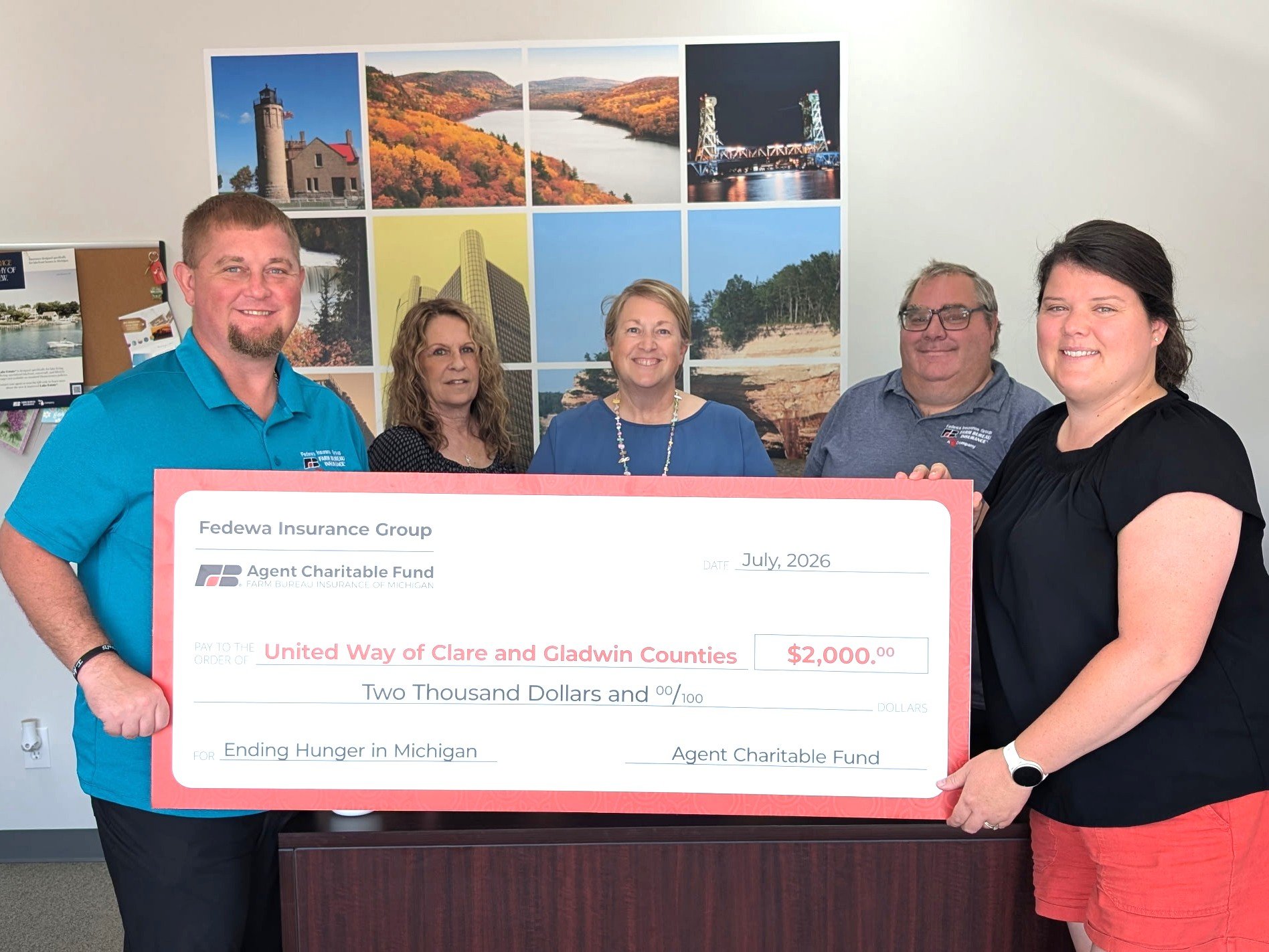 Group of five people holding a large check, standing in front of a wall decorated with scenic photos of lakes, mountains, and cityscapes. The check is made out to United Way of Clare and Gladwin Counties for $2,000, dated July 2026.
