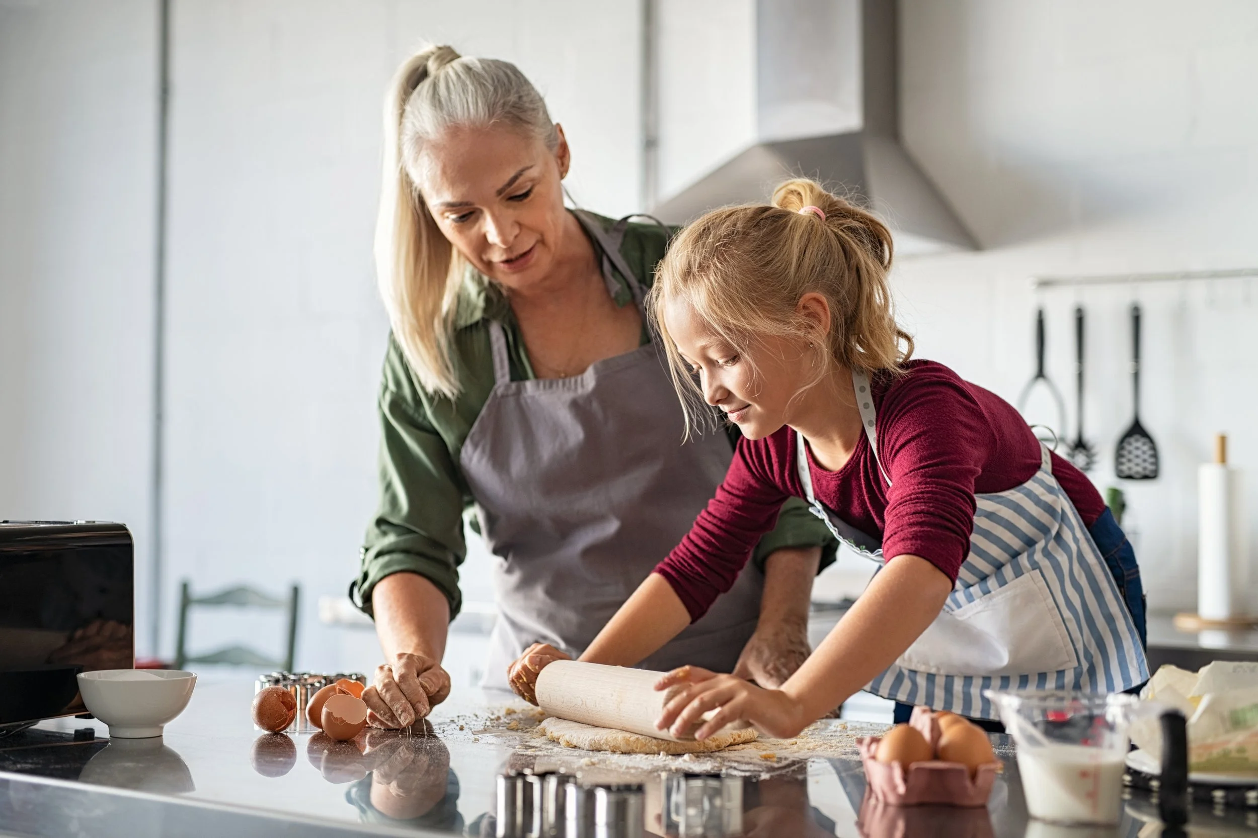 An elderly woman and a young girl baking together in a kitchen. The woman is wearing a gray apron and the girl is in a striped apron. They are rolling dough on a kitchen counter surrounded by eggs, a bowl, and baking tools.
