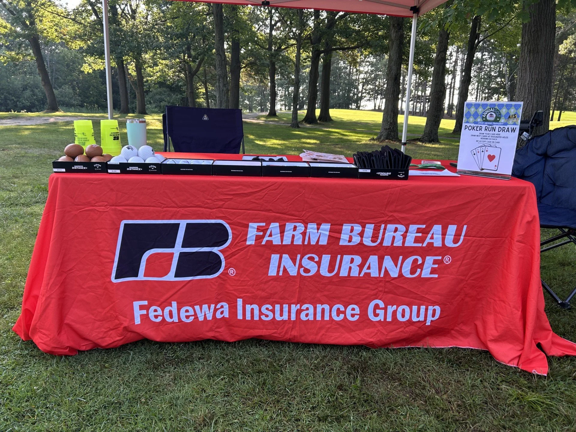 A table covered with a red tablecloth displaying a Federated Insurance Group banner, golf balls, golf tees, and a sign for a poker run draw, set outdoors with trees and grass in the background.