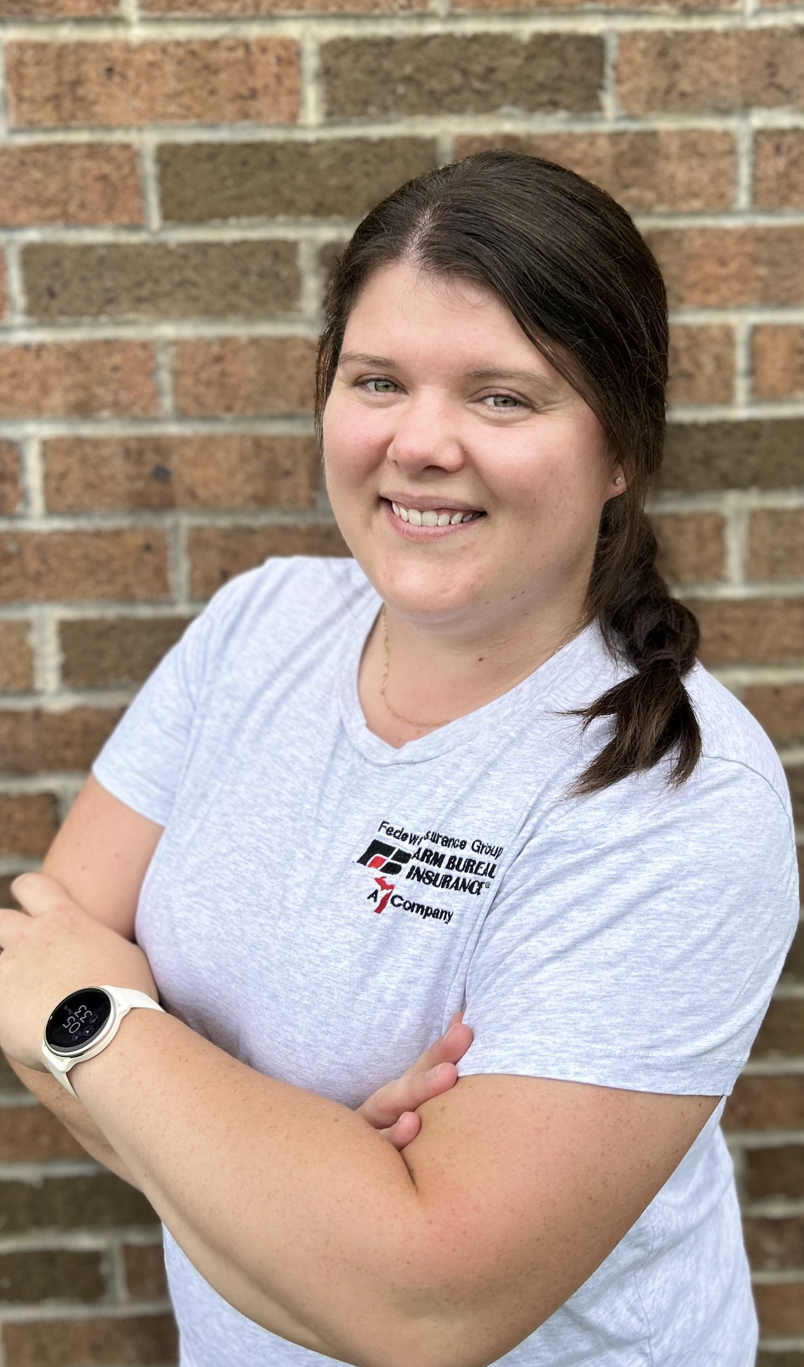 A woman with long brown hair in a side braid, wearing a gray T-shirt with a logo, standing in front of a brick wall, arms crossed and smiling.