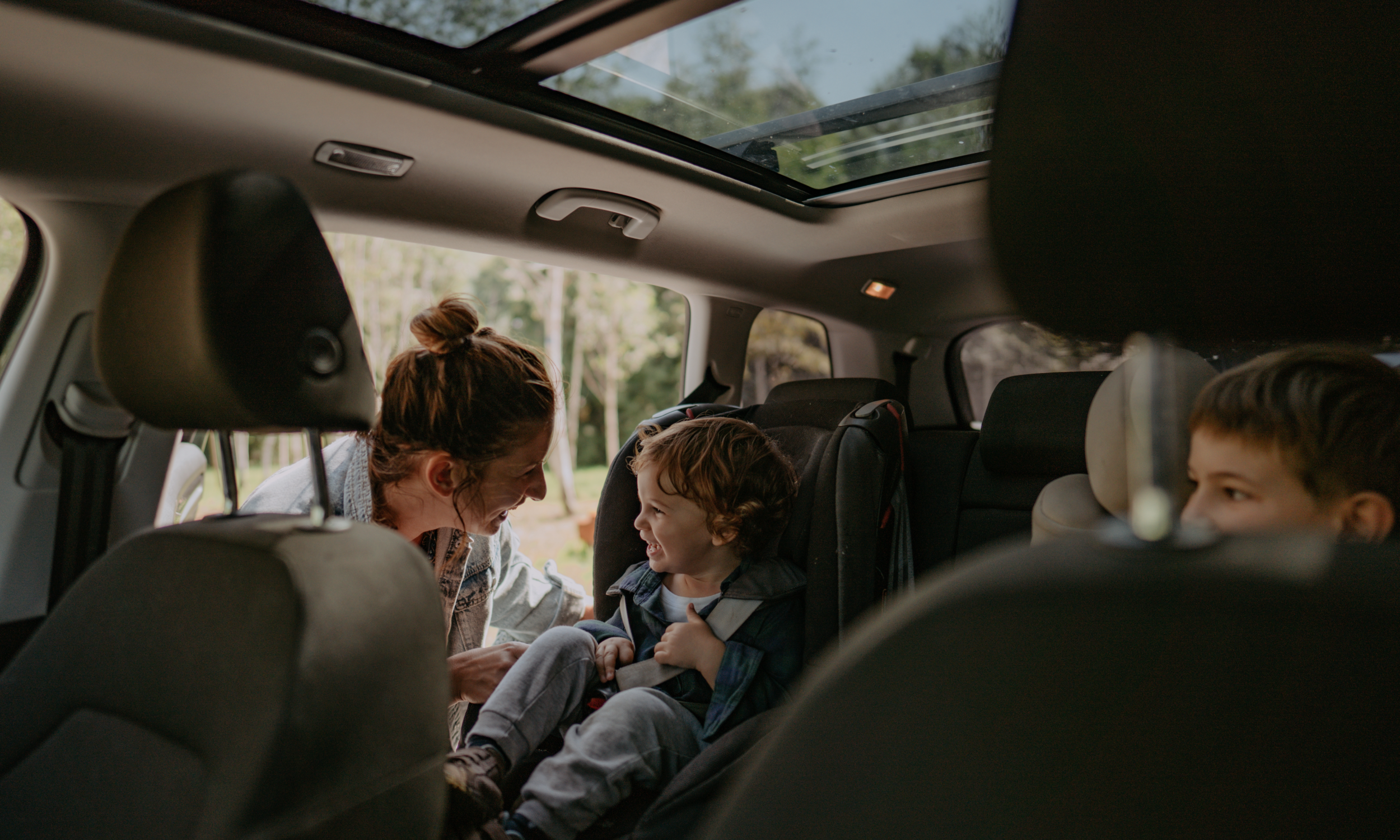 Family of four, including two children and a dog, enjoying a car ride on a sunny day with trees outside the window.