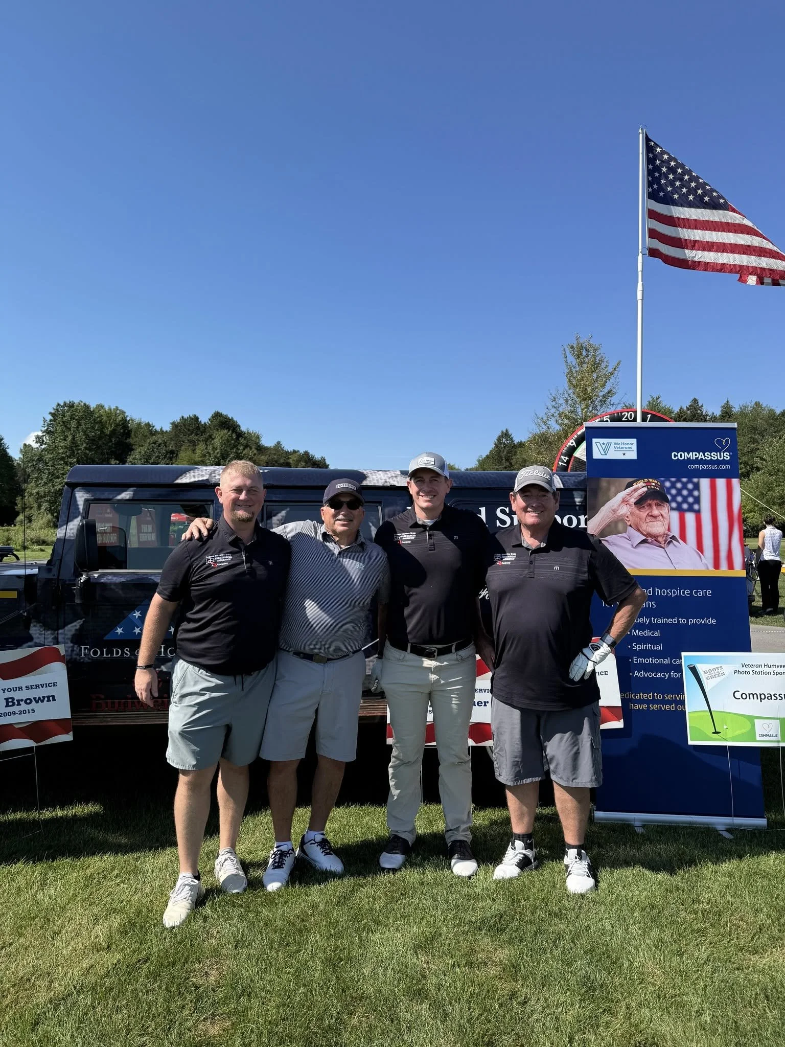 Four men standing together in front of a black vehicle with American flags and signs, outdoors on a sunny day with green grass and trees, smiling at the camera.