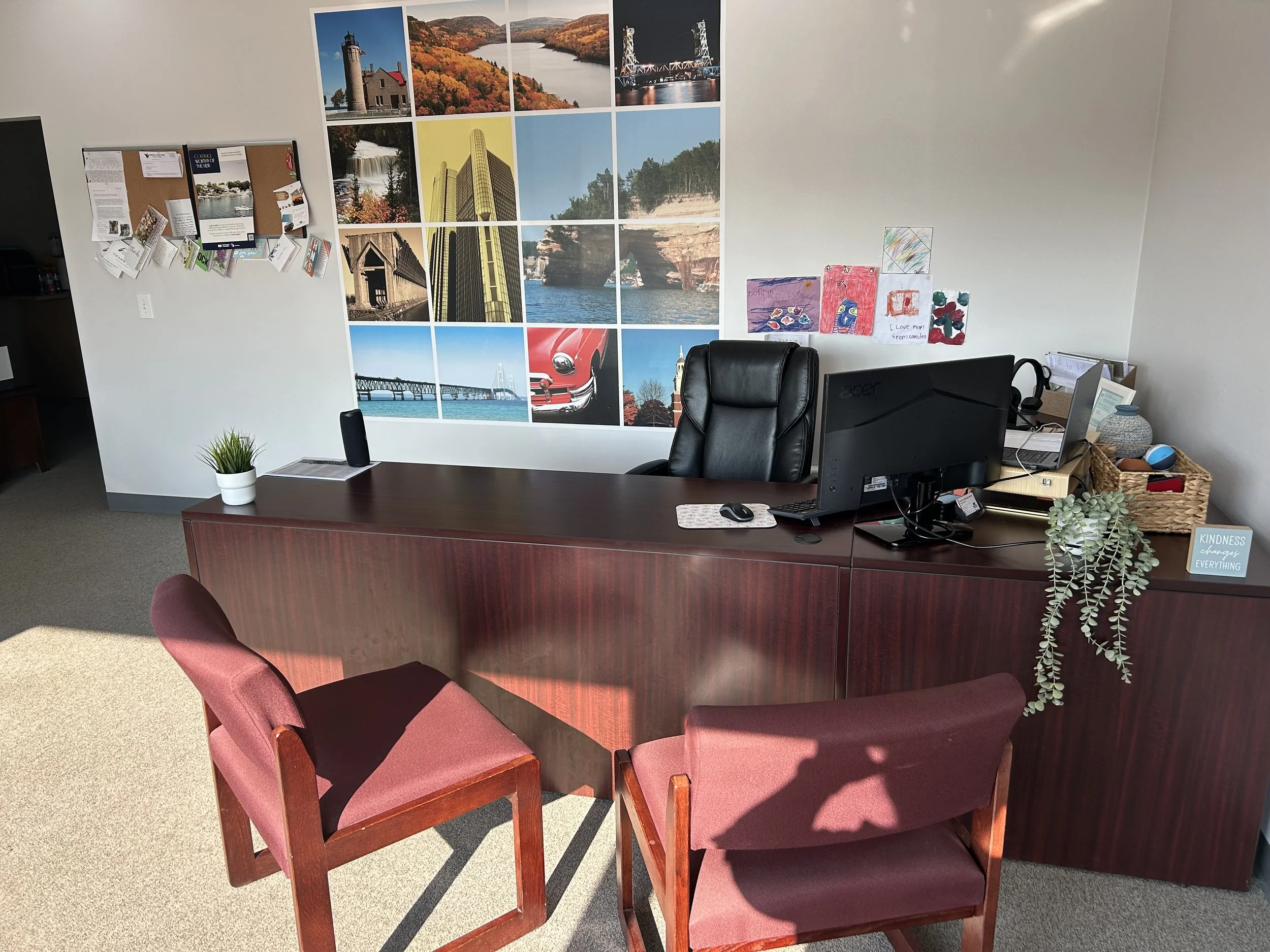 Office workspace with a wooden desk, a computer monitor, a keyboard, a mouse, and a small potted plant. Behind the desk, there are pink cushioned chairs and a wall decorated with a large collage of landscape photographs, including waterfalls, bridges, and cityscapes. There are also children's drawings and notes pinned on a board on the wall.