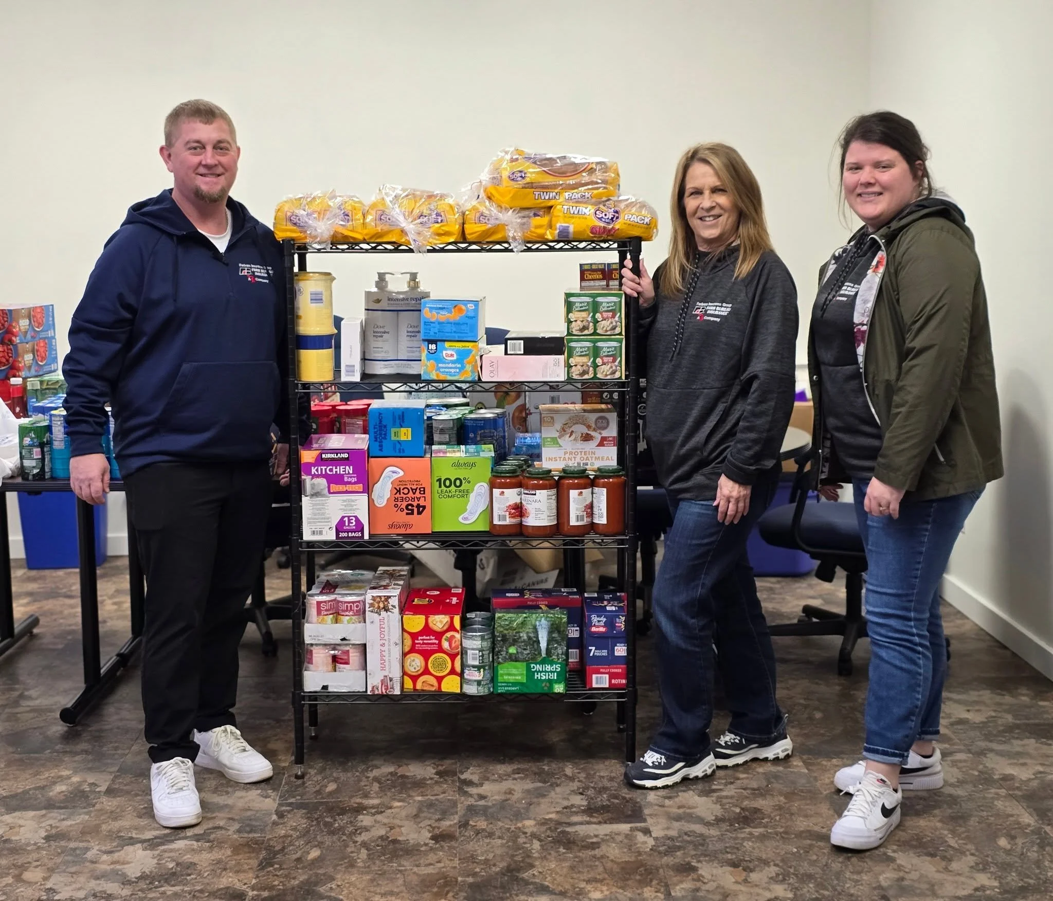 Three people standing behind a black metal shelf stocked with various food and household items, smiling at the camera.