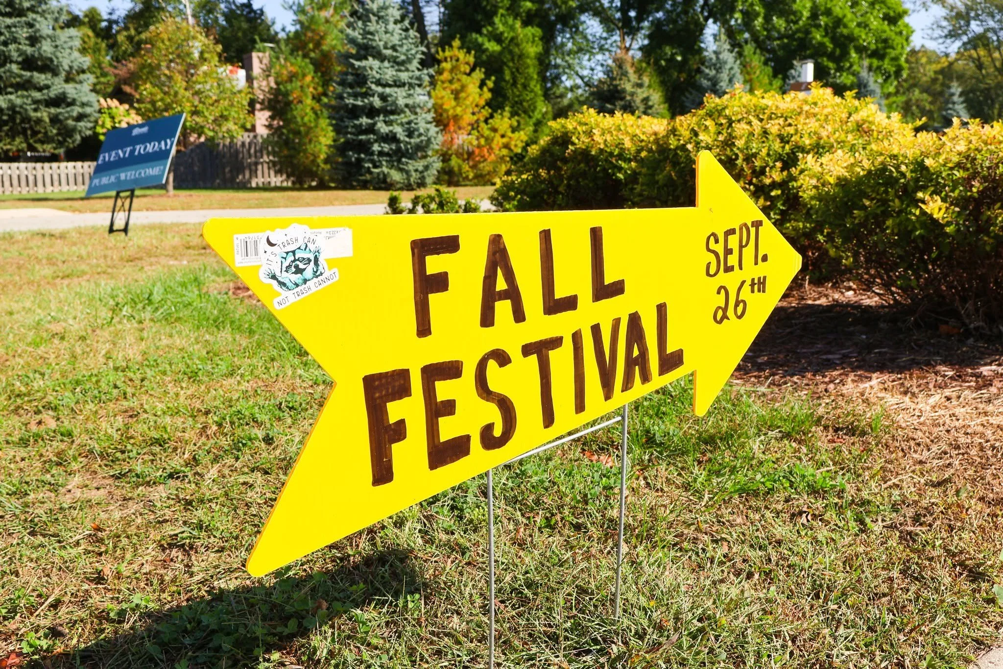 Yellow directional sign for the Fall Festival pointing to September 26th, with a sticker that says 'It's trash can, not trash cannot,' outdoors with trees and bushes in a park or yard.