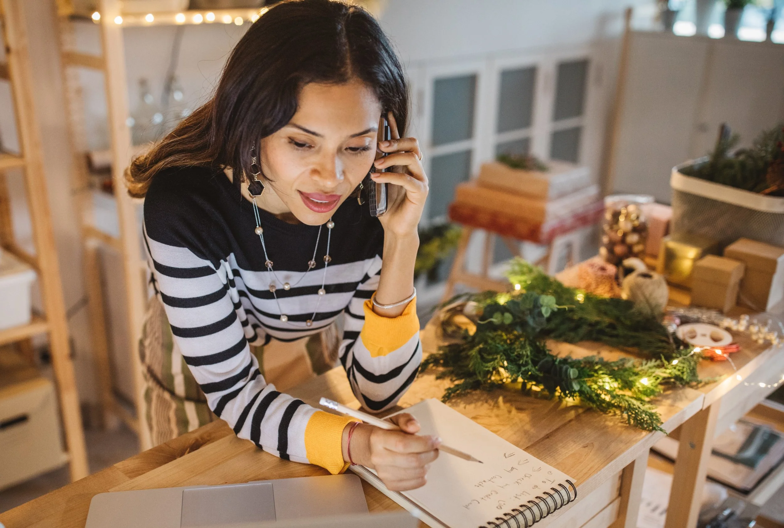 A woman with dark hair, wearing a black and white striped sweater with yellow cuffs, is talking on a phone while writing in a notebook.