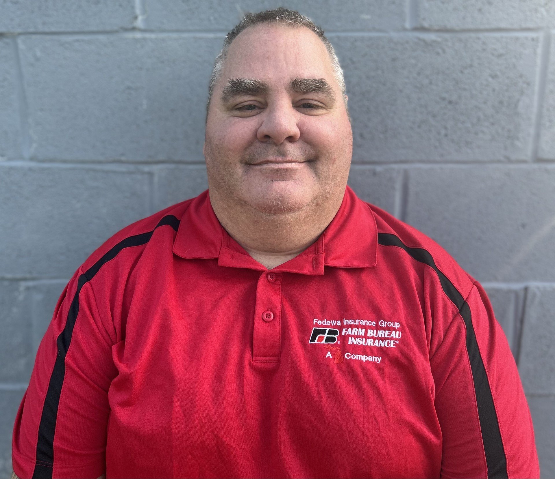 A smiling man wearing a red shirt with black stripes on the sleeves, standing in front of a gray concrete block wall.