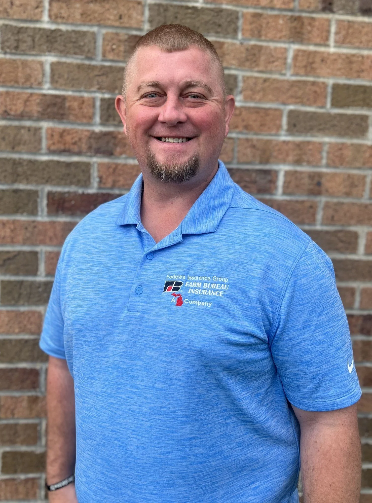 A smiling man with short red hair, facial hair, wearing a blue polo shirt with a logo, standing in front of a brick wall.