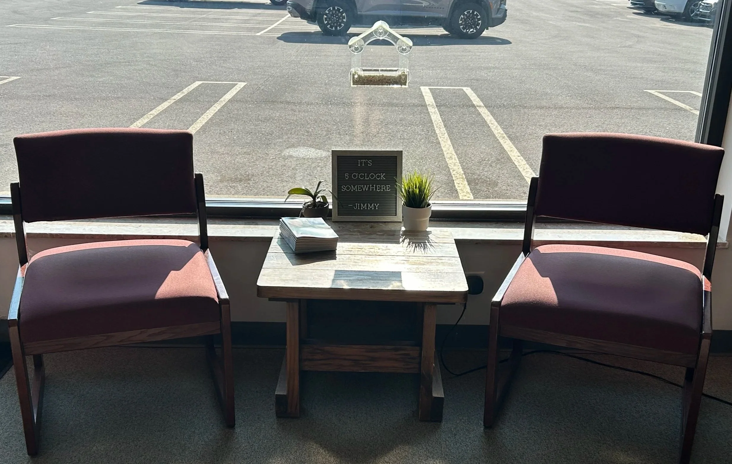 Inside view of a coffee shop or waiting area with two empty chairs facing a large window. On the window sill, there are two small potted plants and a framed sign that reads 'It's 5 o'clock somewhere, Jimmy.' Outside, a parking lot with several cars is visible.