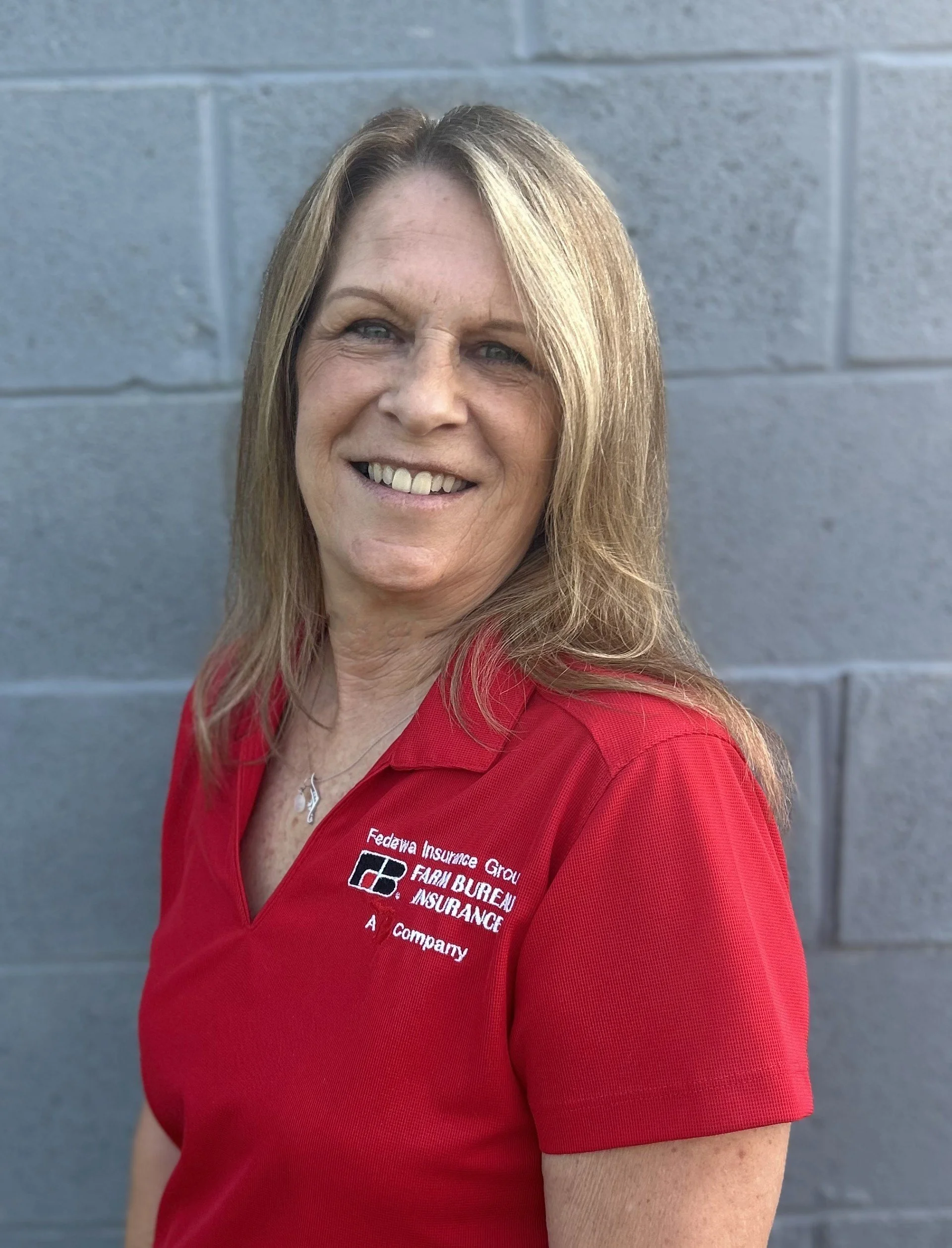 A woman with long blond hair smiling, wearing a red shirt with a logo and text, standing in front of a gray brick wall.