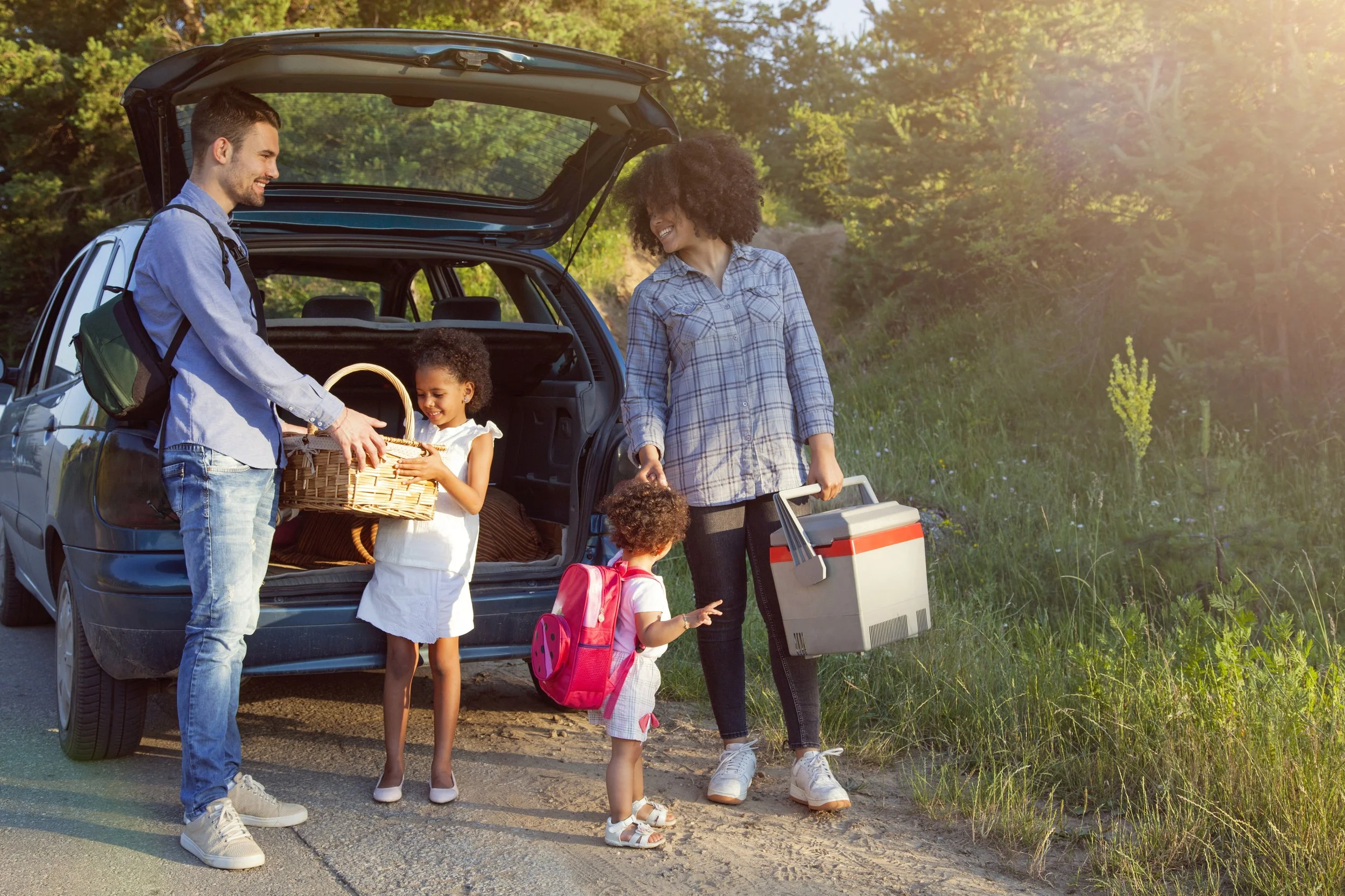 Family loading and unloading items from a car trunk on a sunny day in a forested area, with two children and two adults smiling and engaging in outdoor activity.
