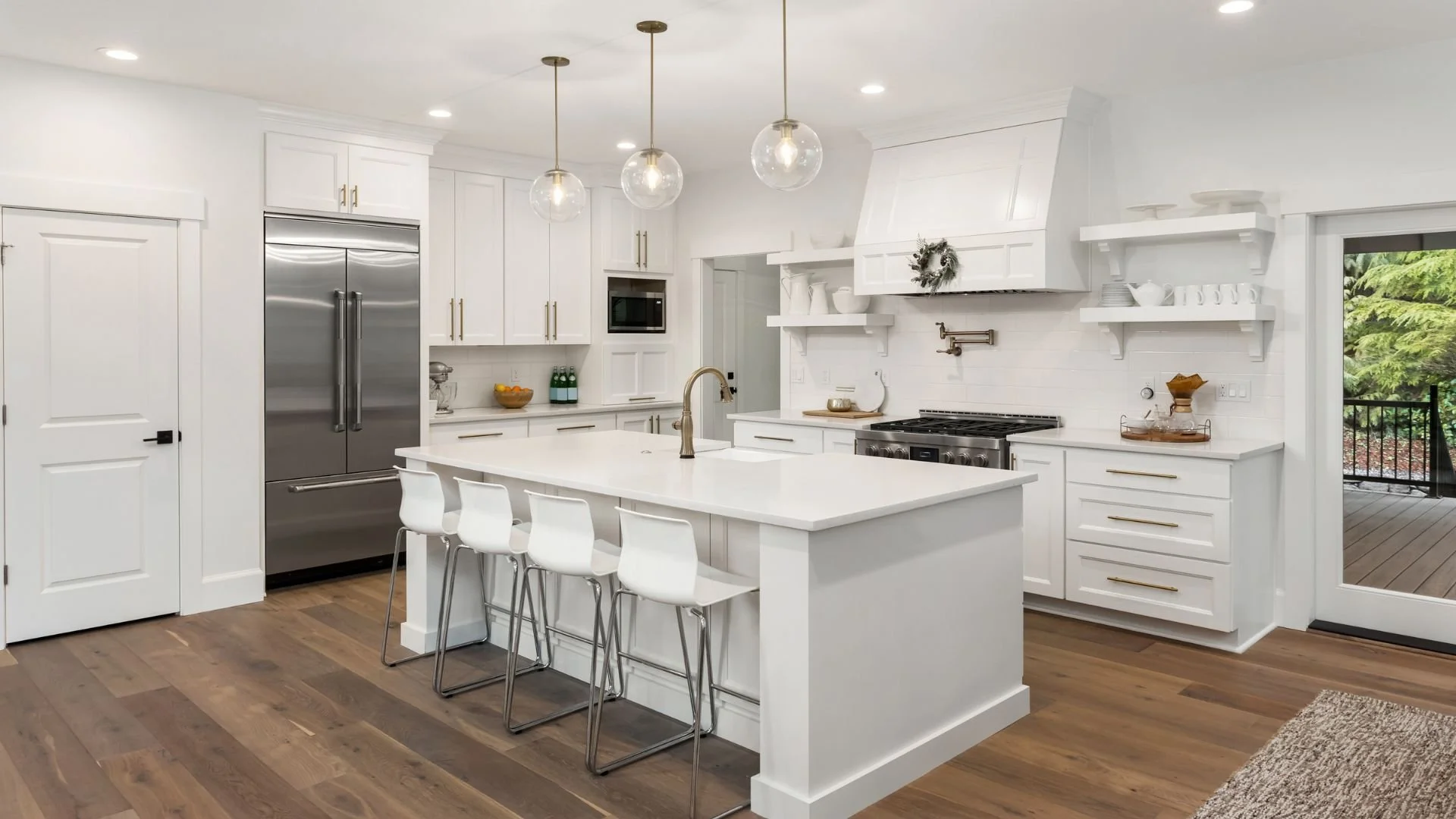 Modern white kitchen with an island, pendant lights, stainless steel appliances, white cabinets, and a sliding door leading to an outdoor deck with trees.