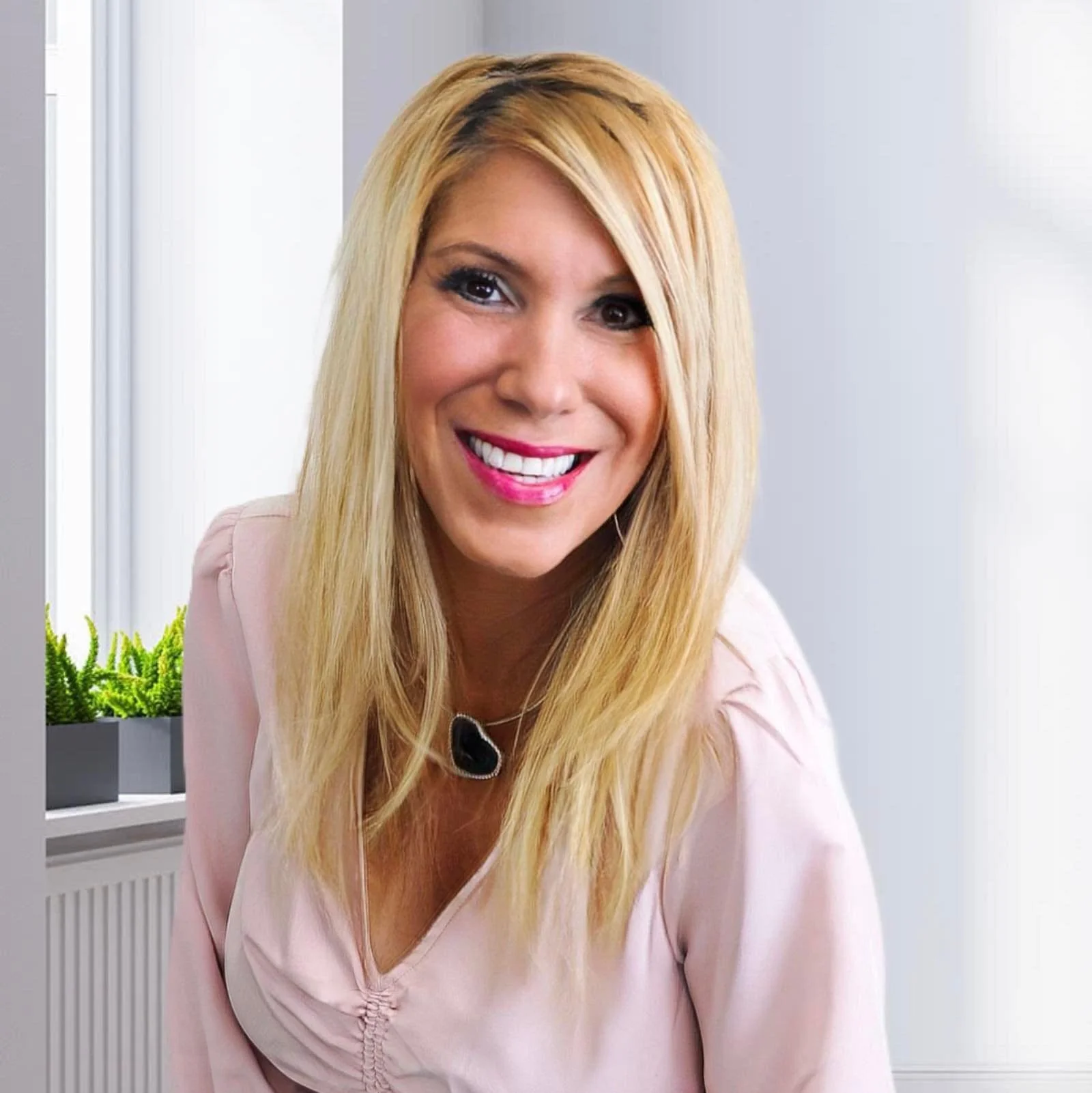 A woman with long blonde hair, wearing a light pink top and a black microphone, smiling in a bright room with plants visible in the background.