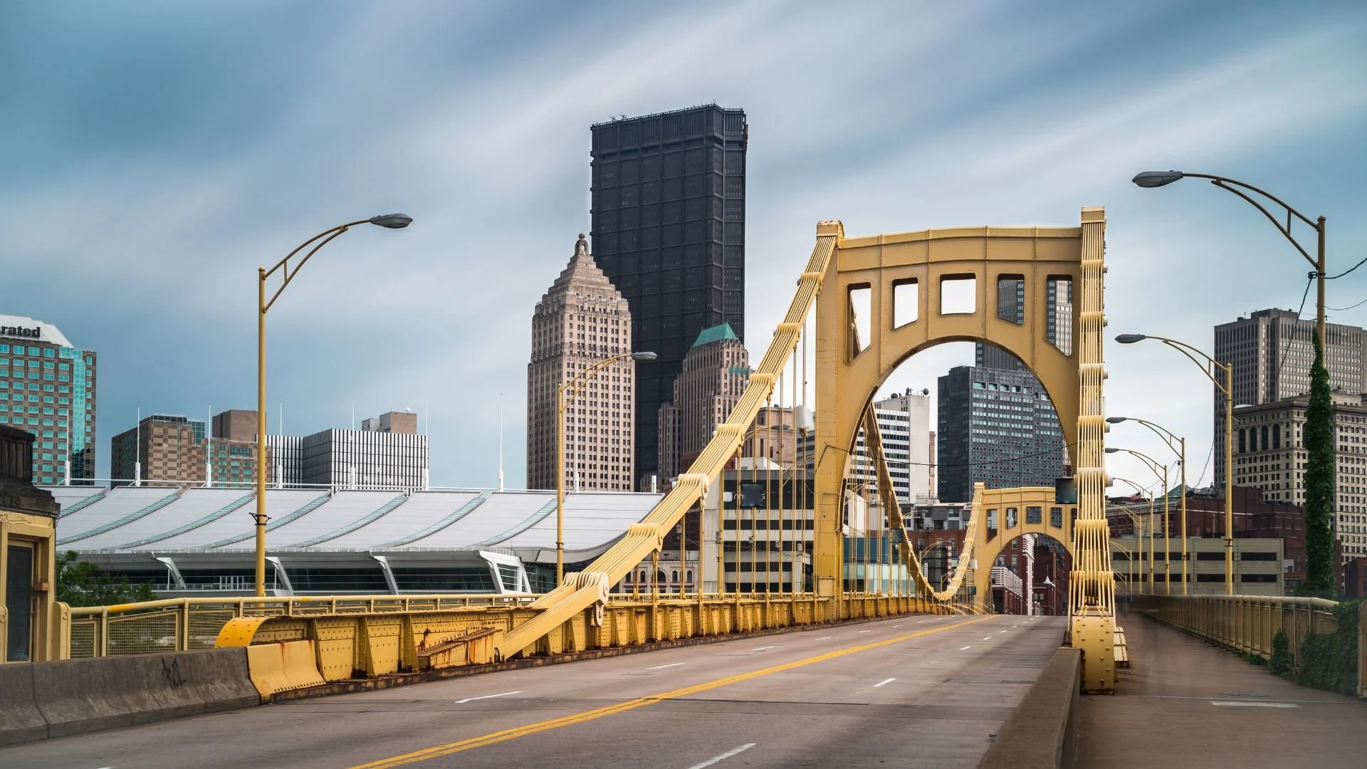 A yellow suspension bridge in an urban city with tall skyscrapers and modern buildings in the background, under a cloudy sky.