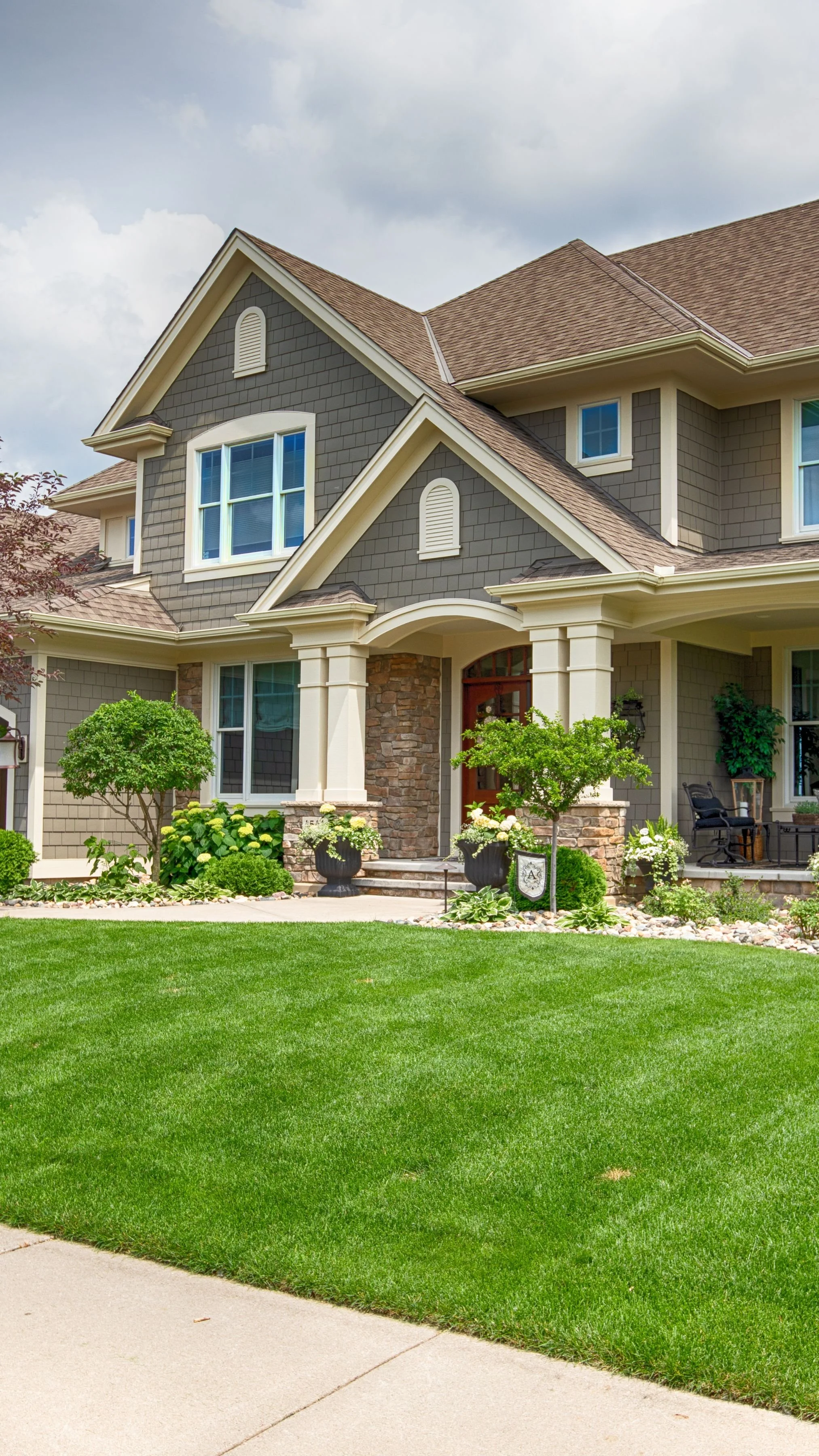 Front yard of a house with a well-maintained green lawn, flowering plants, trees, and a sidewalk.