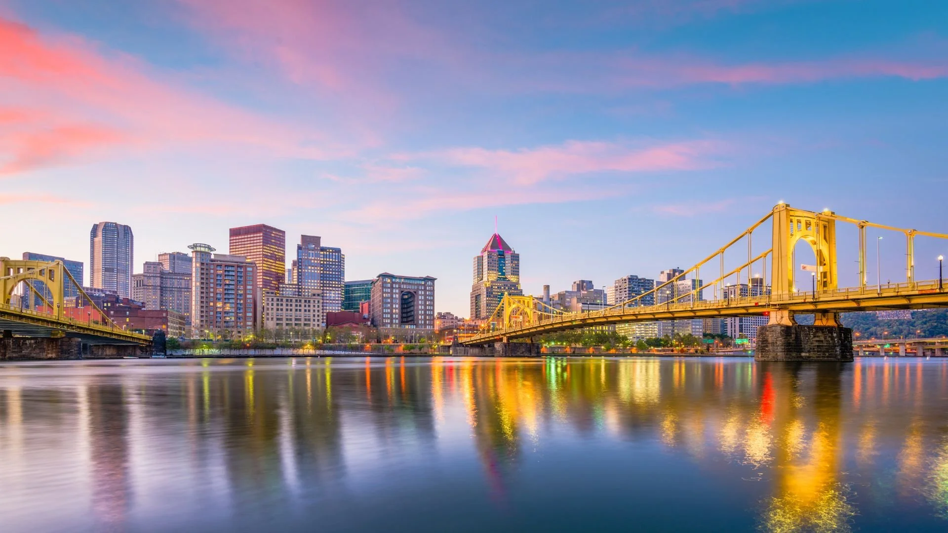 A city skyline with tall buildings, a yellow bridge over a calm river, and a colorful sky at dusk.