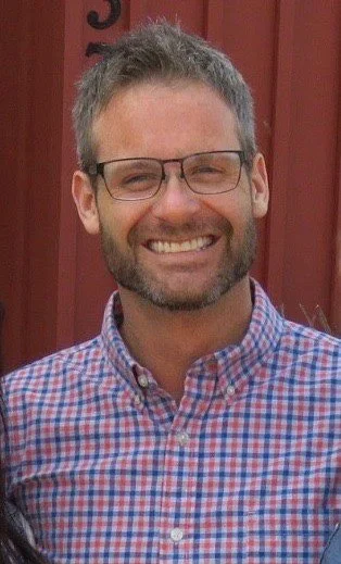 A smiling man with glasses and short gray hair, wearing a checkered shirt, standing in front of a red wooden background.