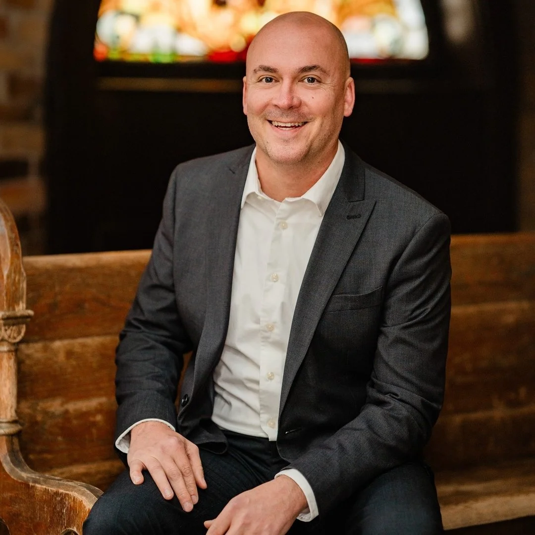 Smiling man in gray suit and white shirt sitting on a wooden bench in a cozy indoor setting.