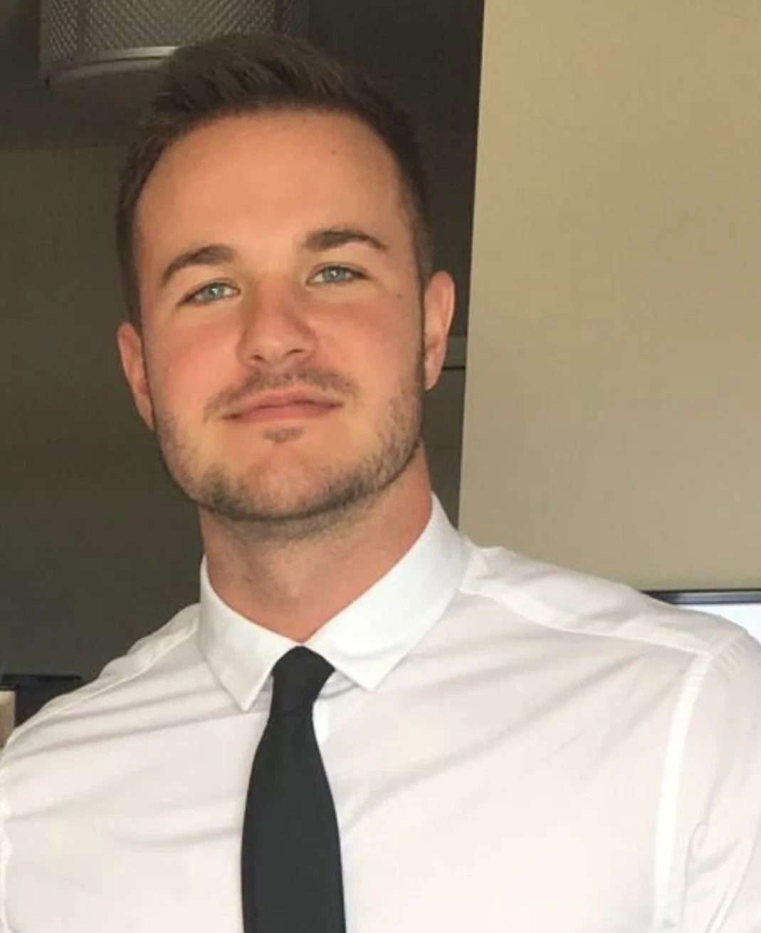A young man with short brown hair, blue eyes, and a slight beard, wearing a white dress shirt and a black tie, smiling indoors.
