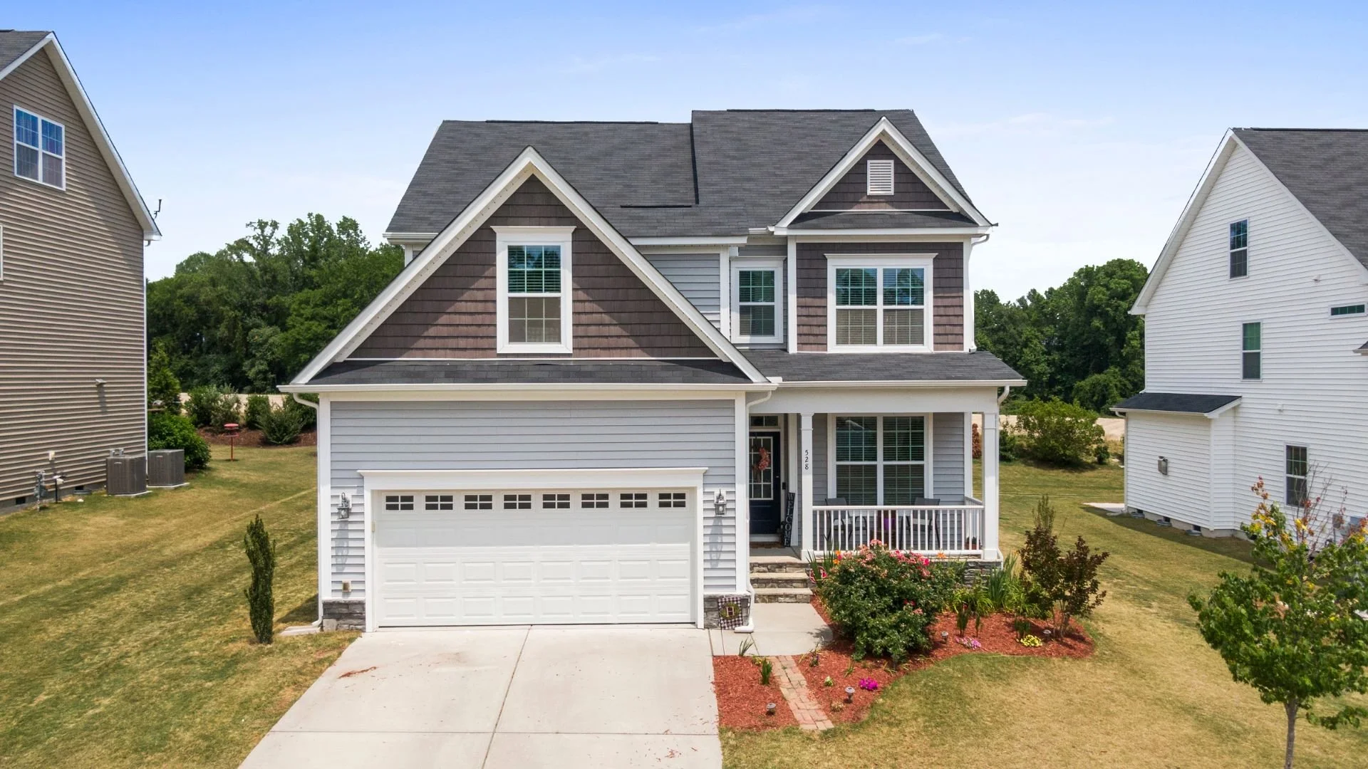 Two-story suburban house with gray and white siding, front porch with white railing, and a two-car garage. Well-kept front yard with flowers and shrubs, neighboring houses on each side, and a green backyard with trees.