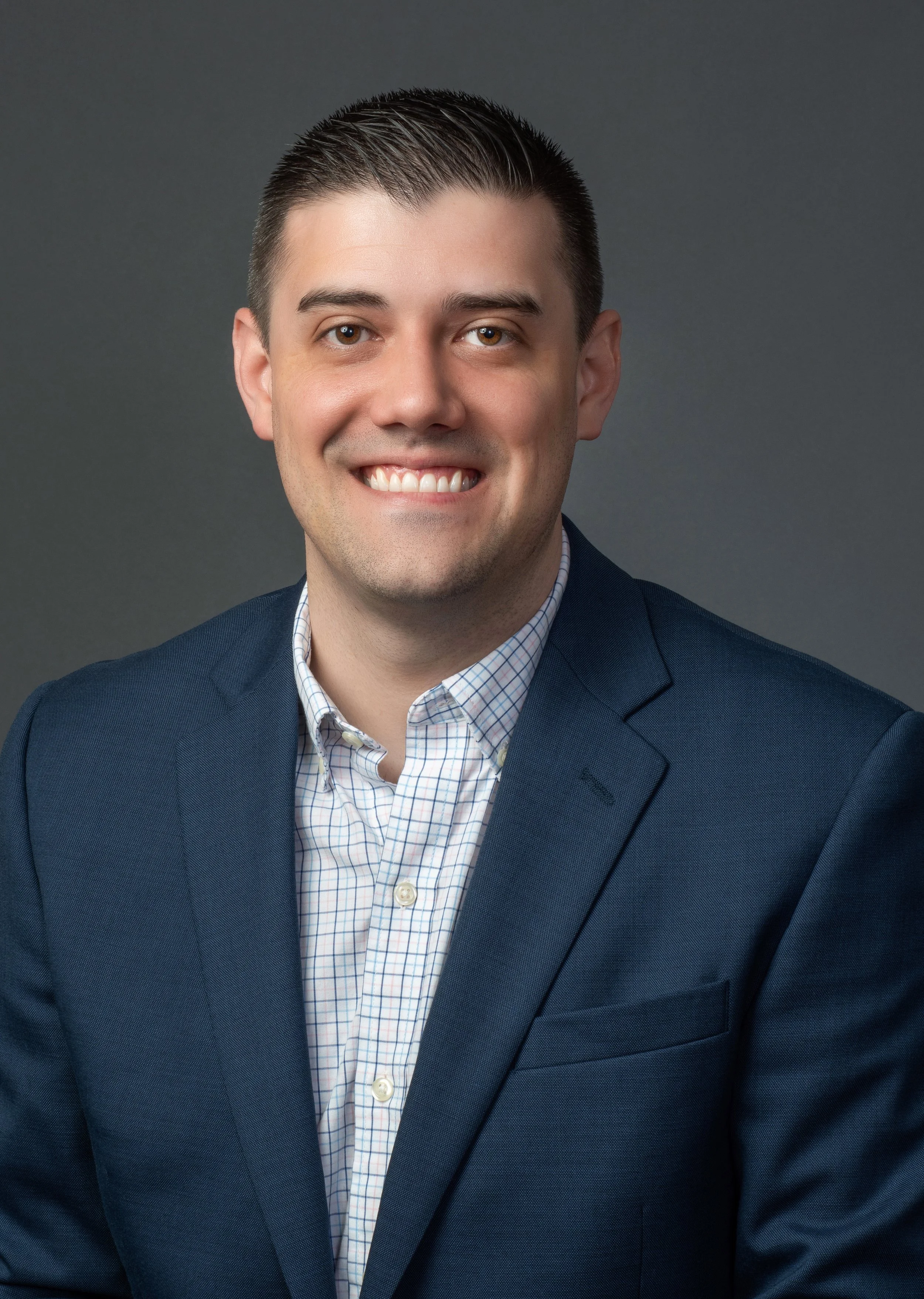 Portrait of a smiling man with dark hair wearing a navy blazer, light checkered shirt, against a gray background.