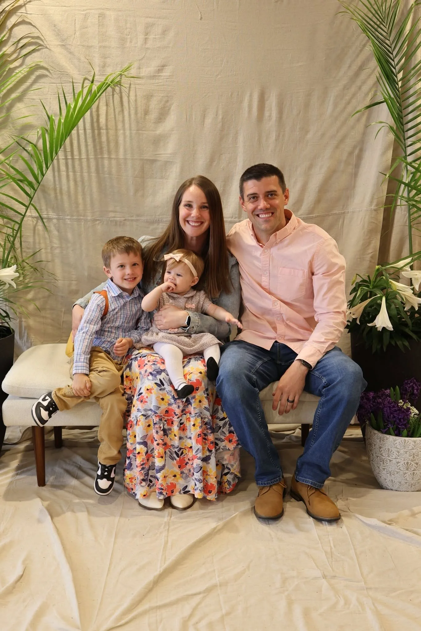 A family of five sitting on a bench in front of a beige curtain with green plants on both sides. The family includes a man, woman, and three children, all smiling at the camera.