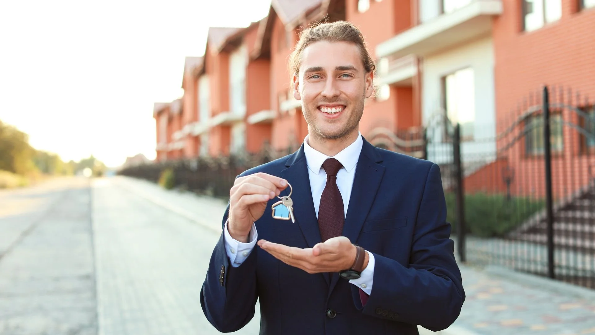 A man in a business suit smiling and holding a set of house keys with a house-shaped keychain.