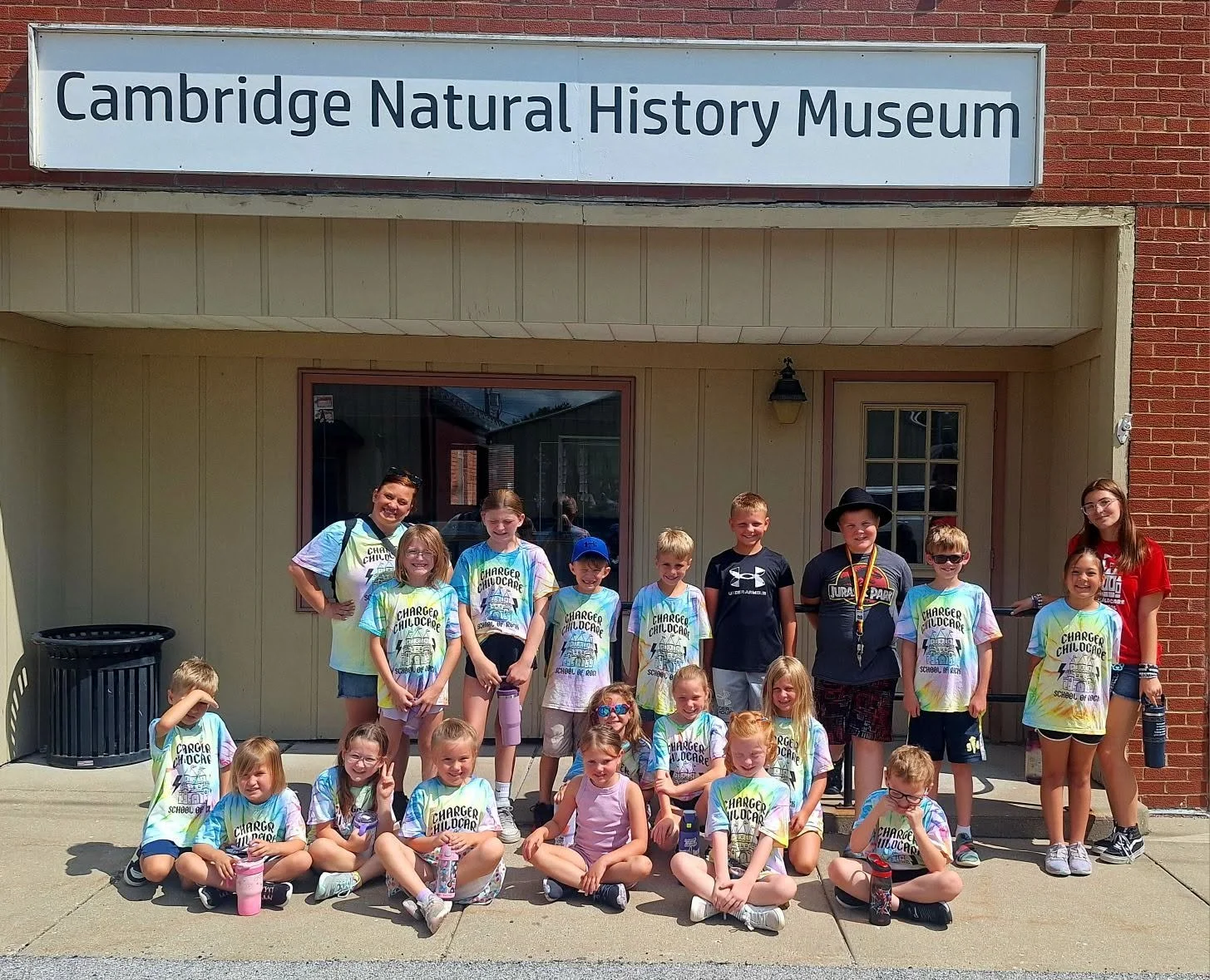 Young visitors and adult chaperones pose together outside Cambridge Natural History Museum after a group visit.