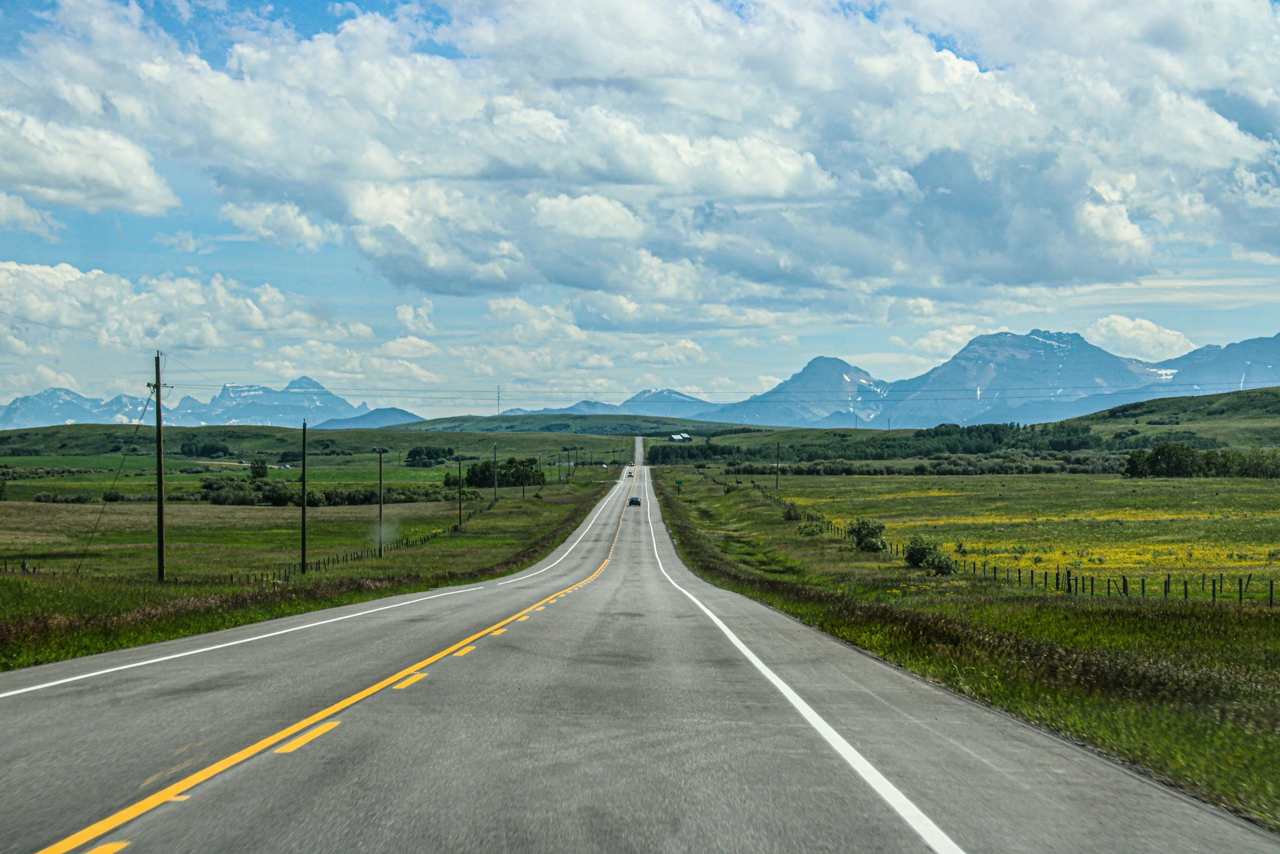 Long straight road through green countryside with mountains in the background under a partly cloudy sky.