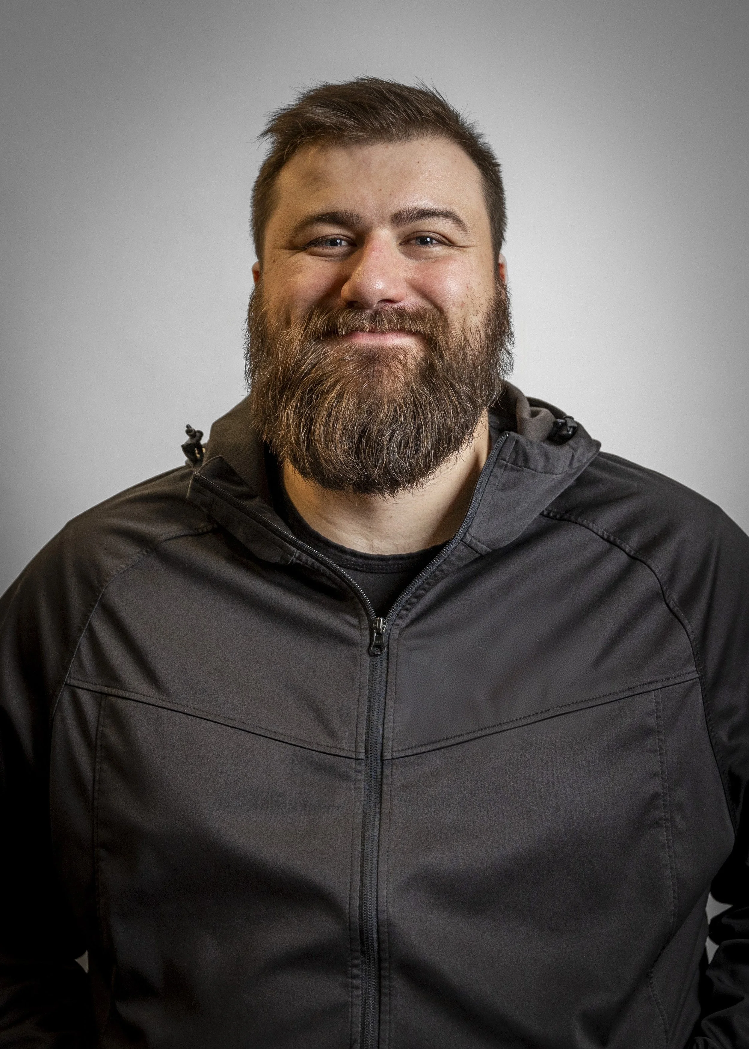 A man with a full beard and short hair wearing a black jacket, smiling at the camera against a plain gray background.