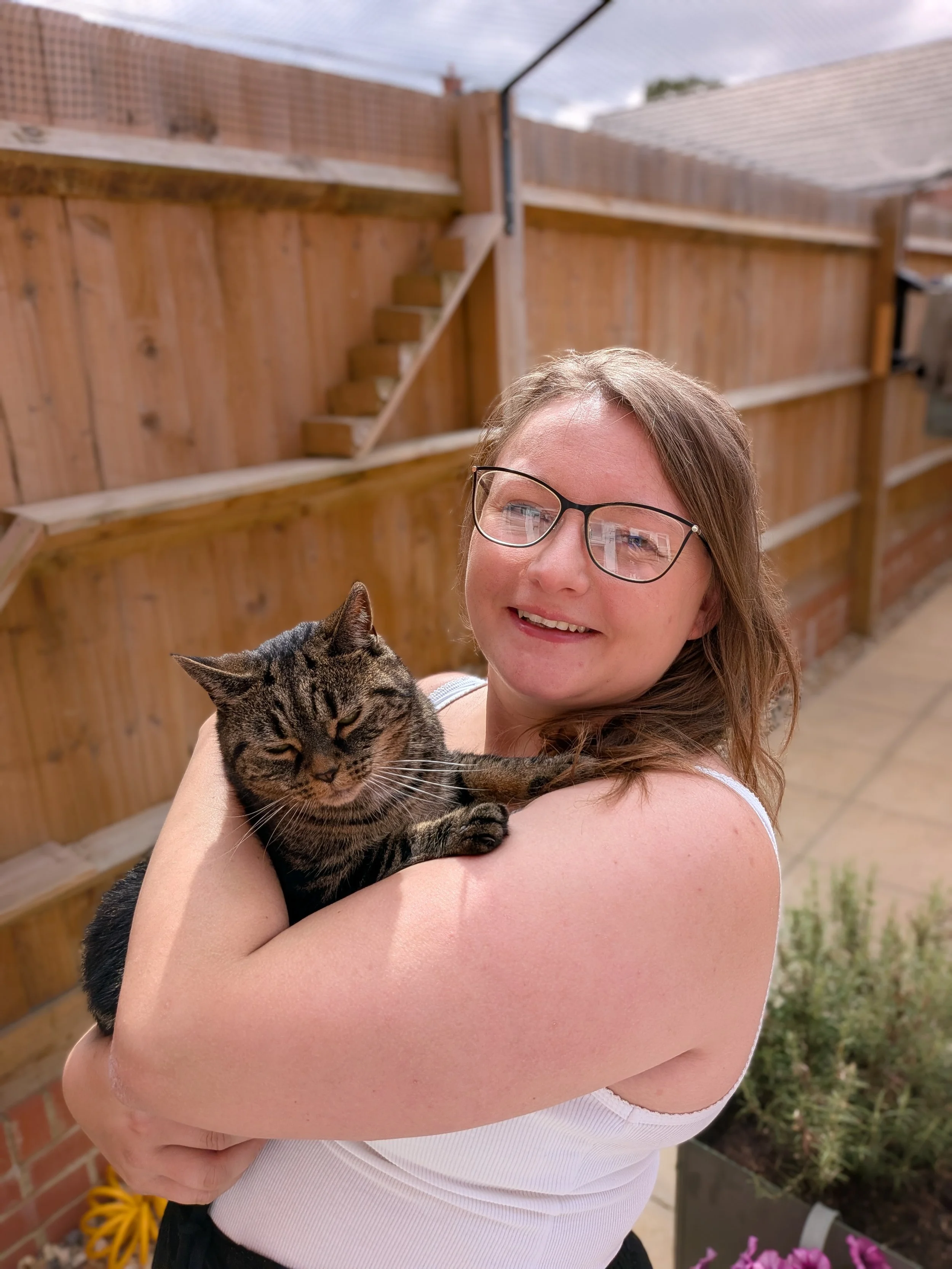 A woman with glasses holding a tabby cat outside in a backyard with a wooden fence and potted plants.
