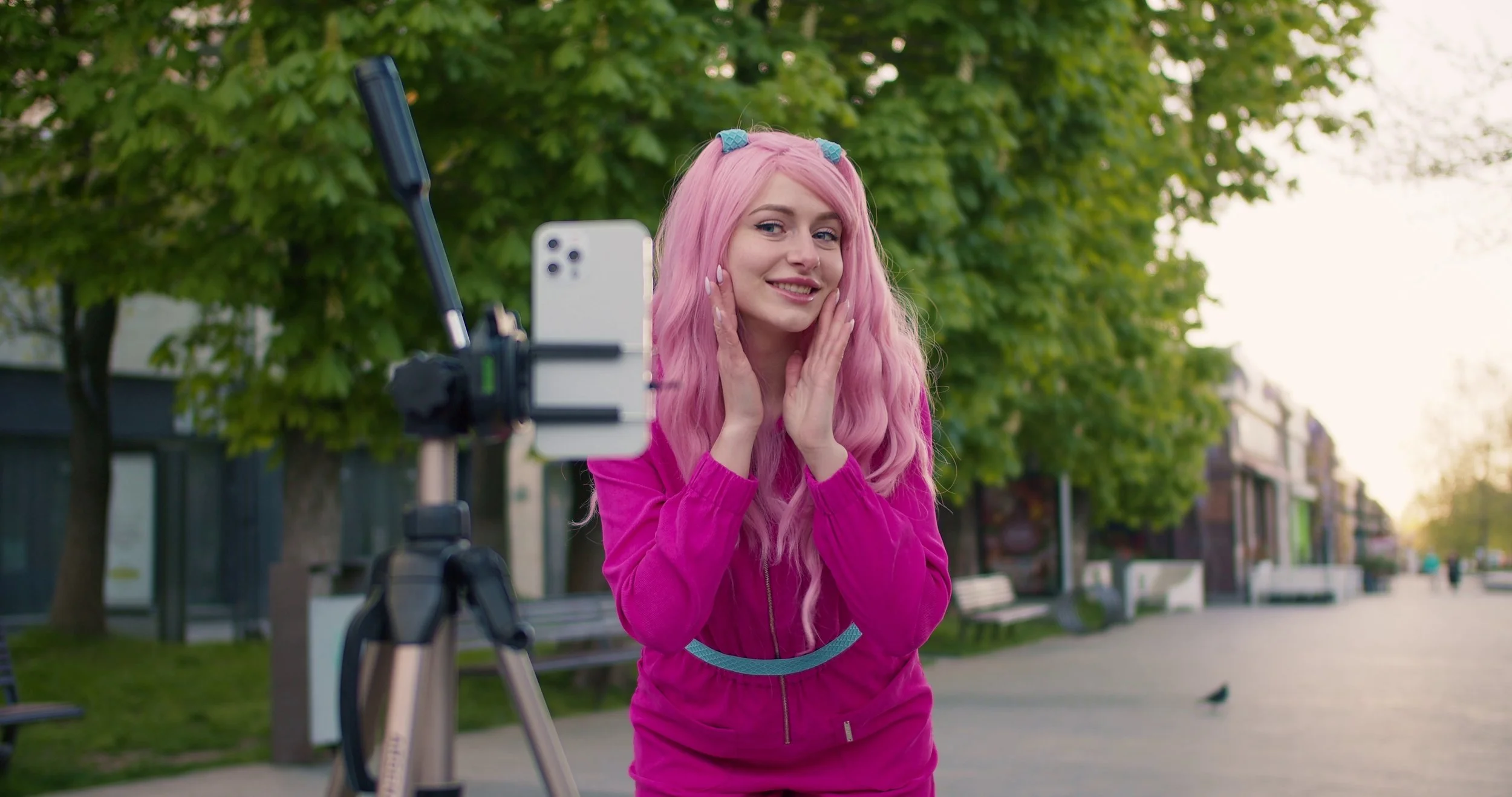 Young woman with pink hair in a pink jacket, smiling and posing as she looks at her reflection in a smartphone mounted on a tripod, outdoors in a park with green trees.