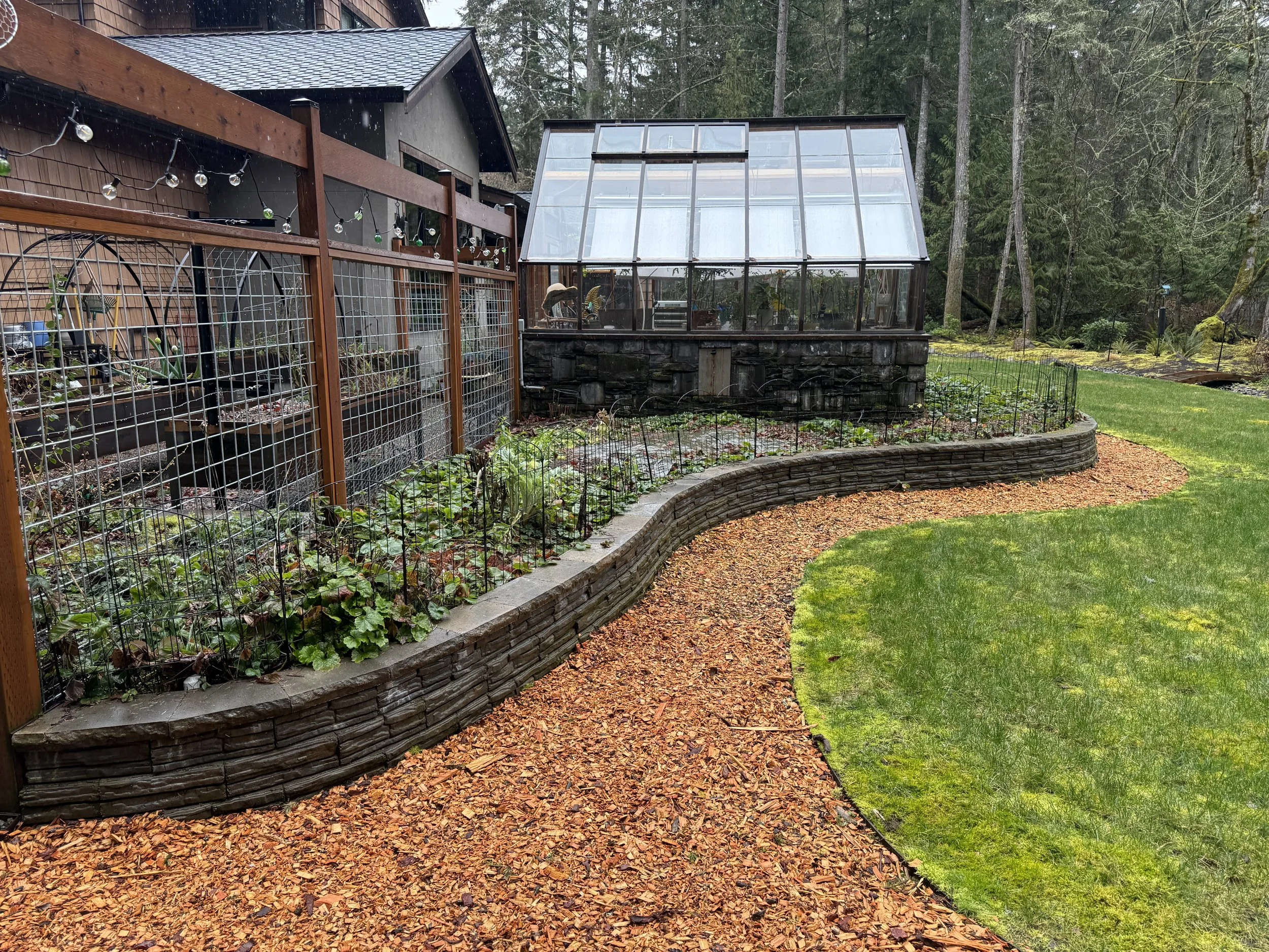 A backyard garden with a greenhouse on a stone foundation, surrounded by wooden fencing and lush green trees, with a curved brick-lined garden bed and mulch path.
