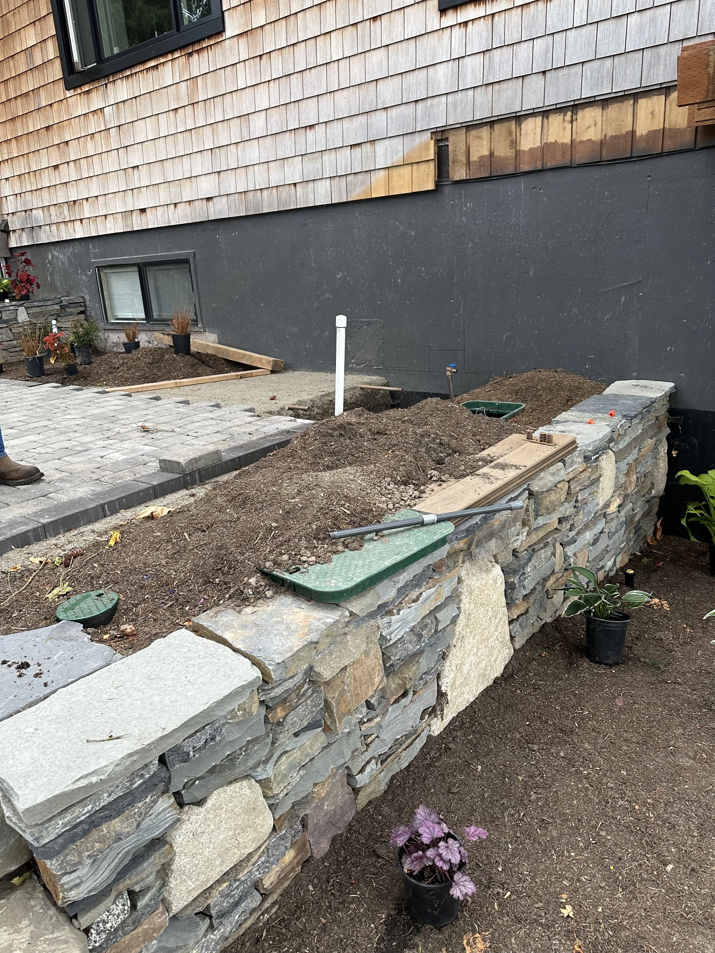 A garden area under construction with a stone retaining wall, potted plants, dirt, and a partially completed paved pathway next to a house with wooden siding.